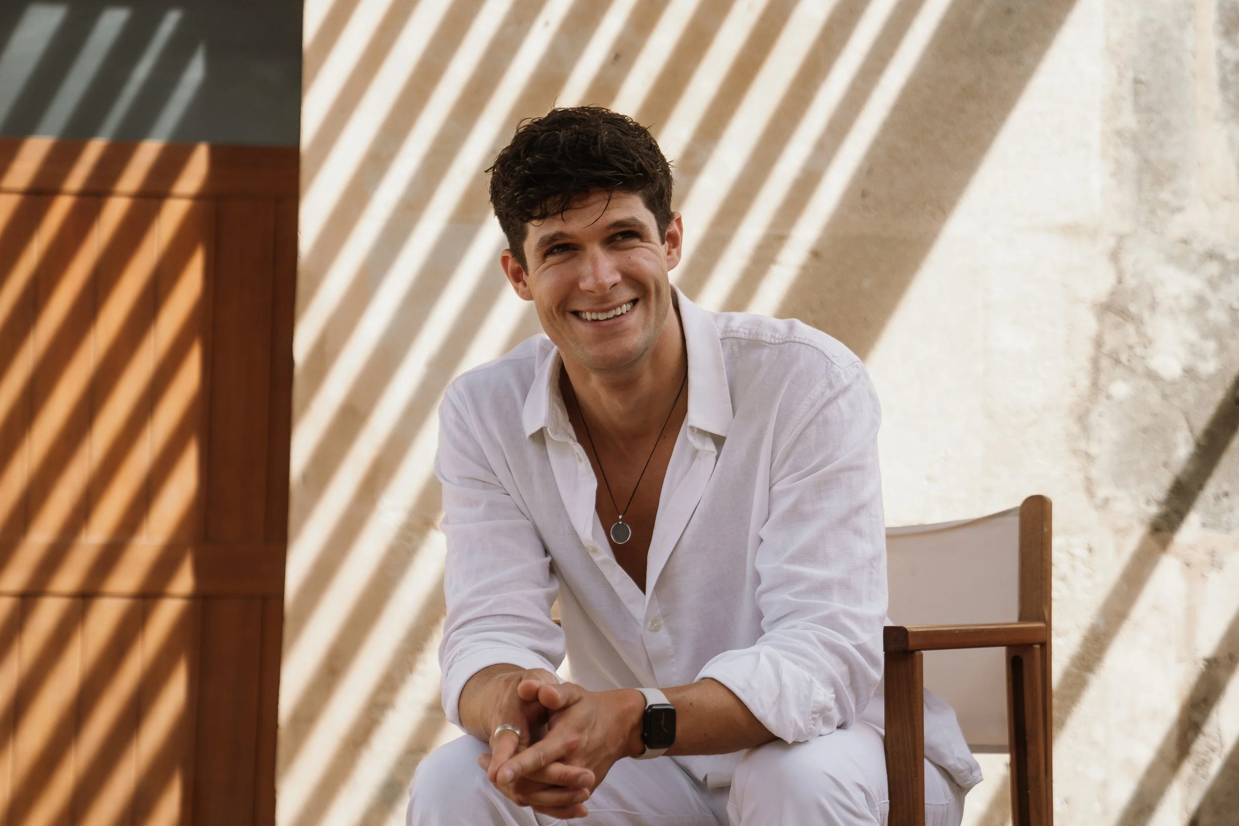 Smiling man in white shirt seated, with striped shadows on a wall behind him.