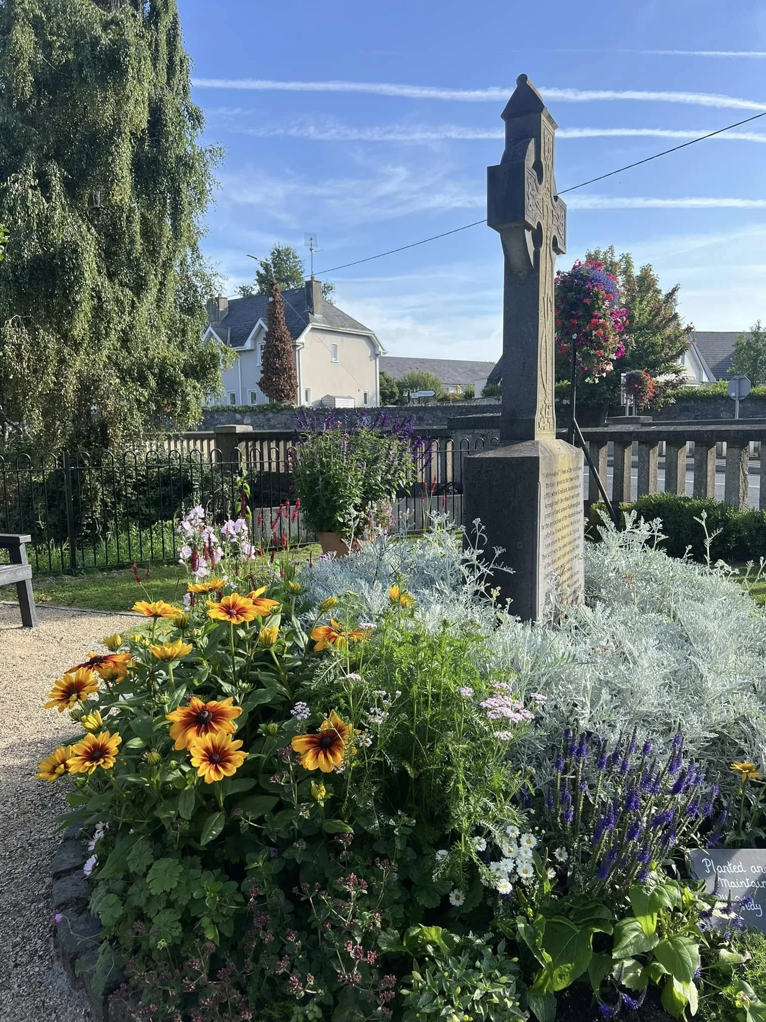 A garden with colorful flowers and greenery surrounding a stone cross monument in a small town setting, under a clear blue sky.