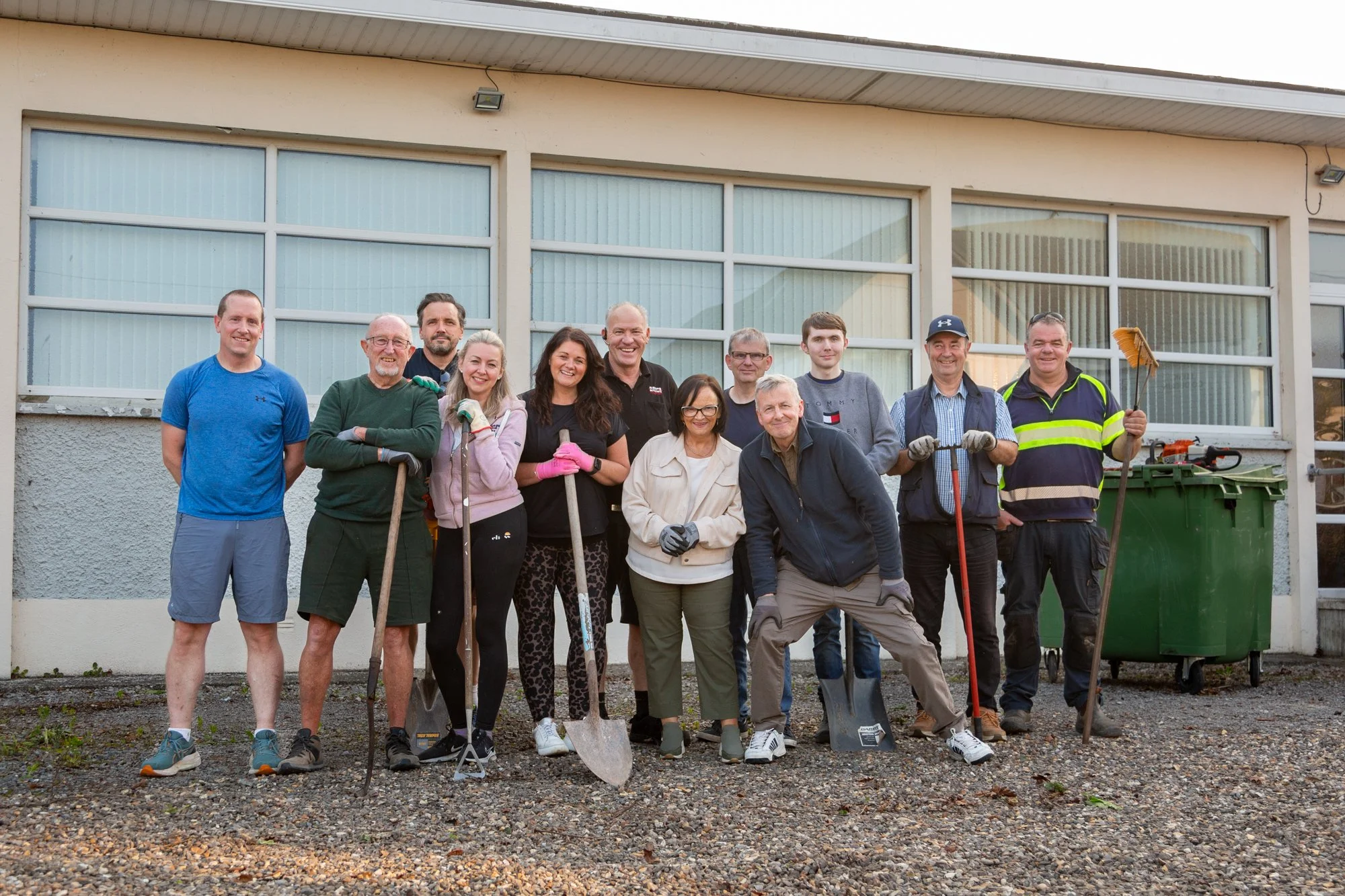 Group of people standing outside in front of a building, holding gardening tools, smiling, dressed casually for a community cleanup or gardening event.