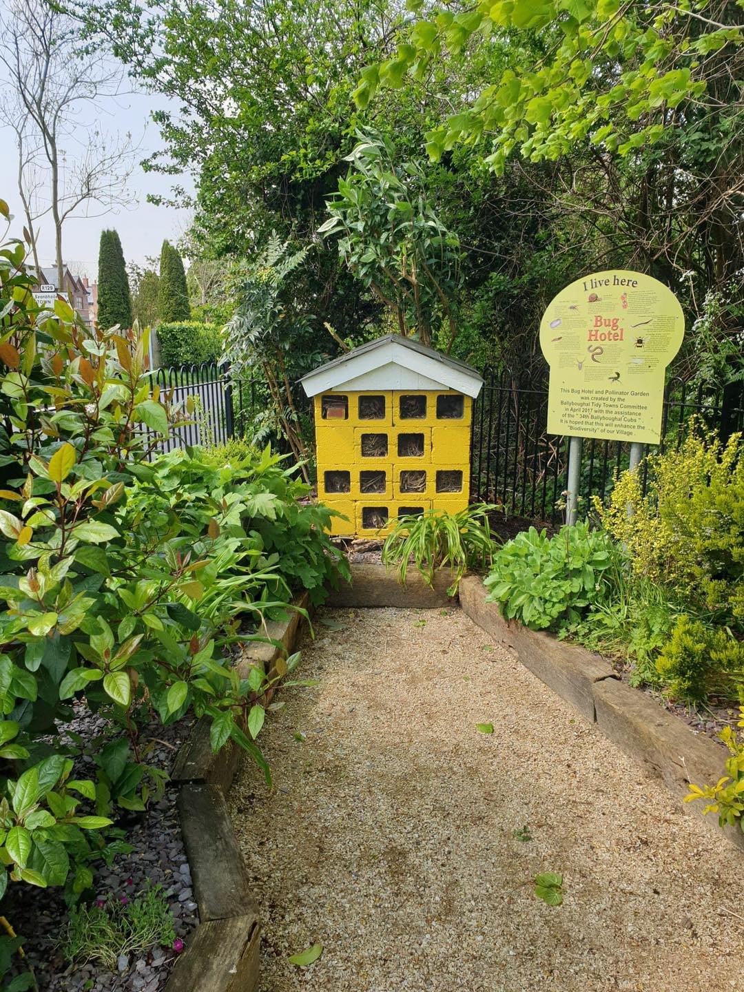 Small yellow insect hotel with a white roof, surrounded by green plants and shrubs, located in a garden with a gravel pathway and a sign that reads 'I live here' and explains it's a bug hotel and pollinator garden.