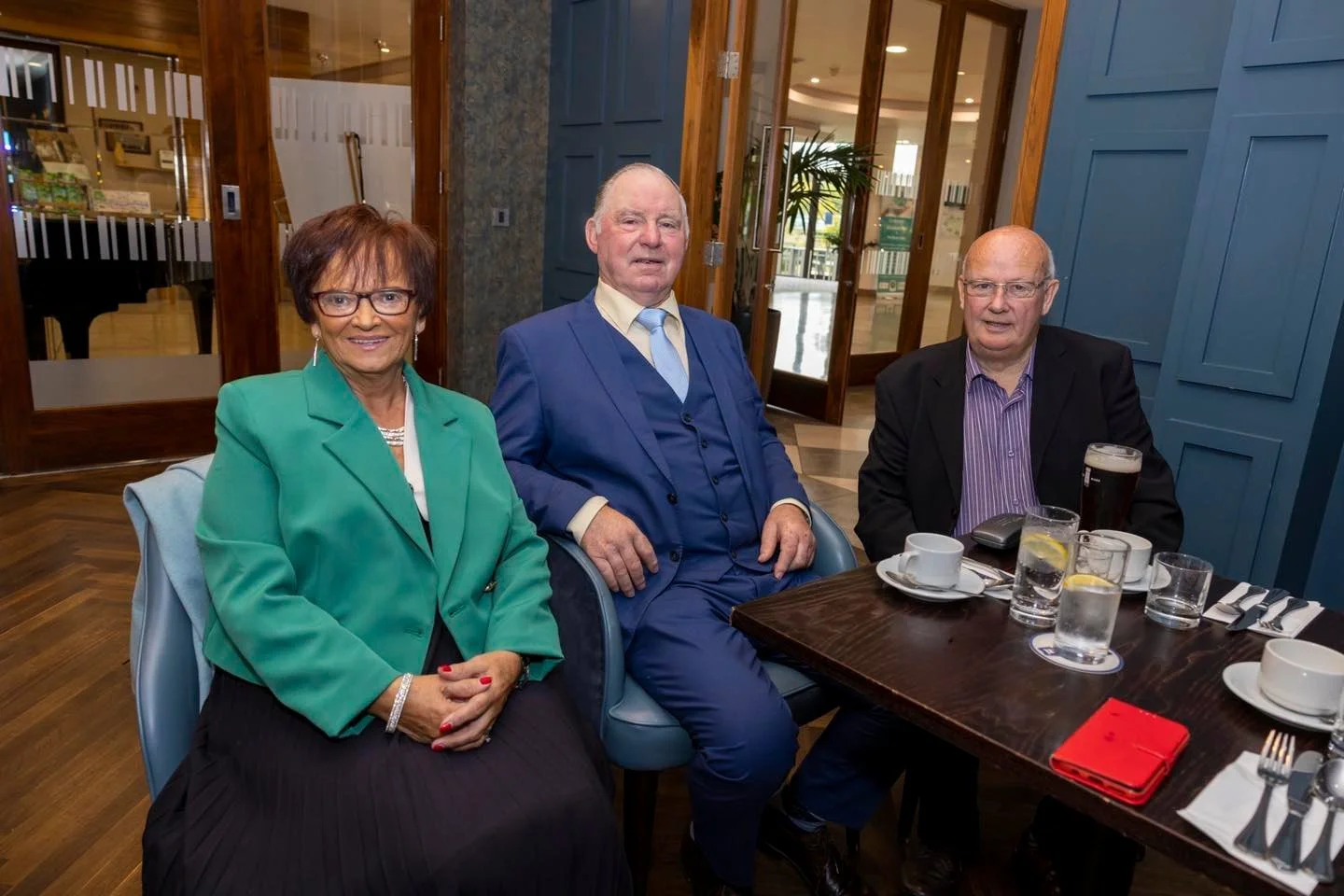 Three seniors sitting at a dining table in a restaurant, smiling at the camera. The woman on the left wears a green blazer, the man in the middle wears a blue suit, and the man on the right wears a black blazer with a purple shirt. The table has drin