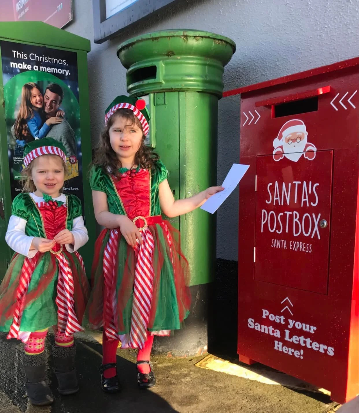 Two young girls dressed as elves with red, green, and white costumes, standing next to a green mailbox and a red Santa mailbox. One girl is holding a letter, ready to mail it to Santa.
