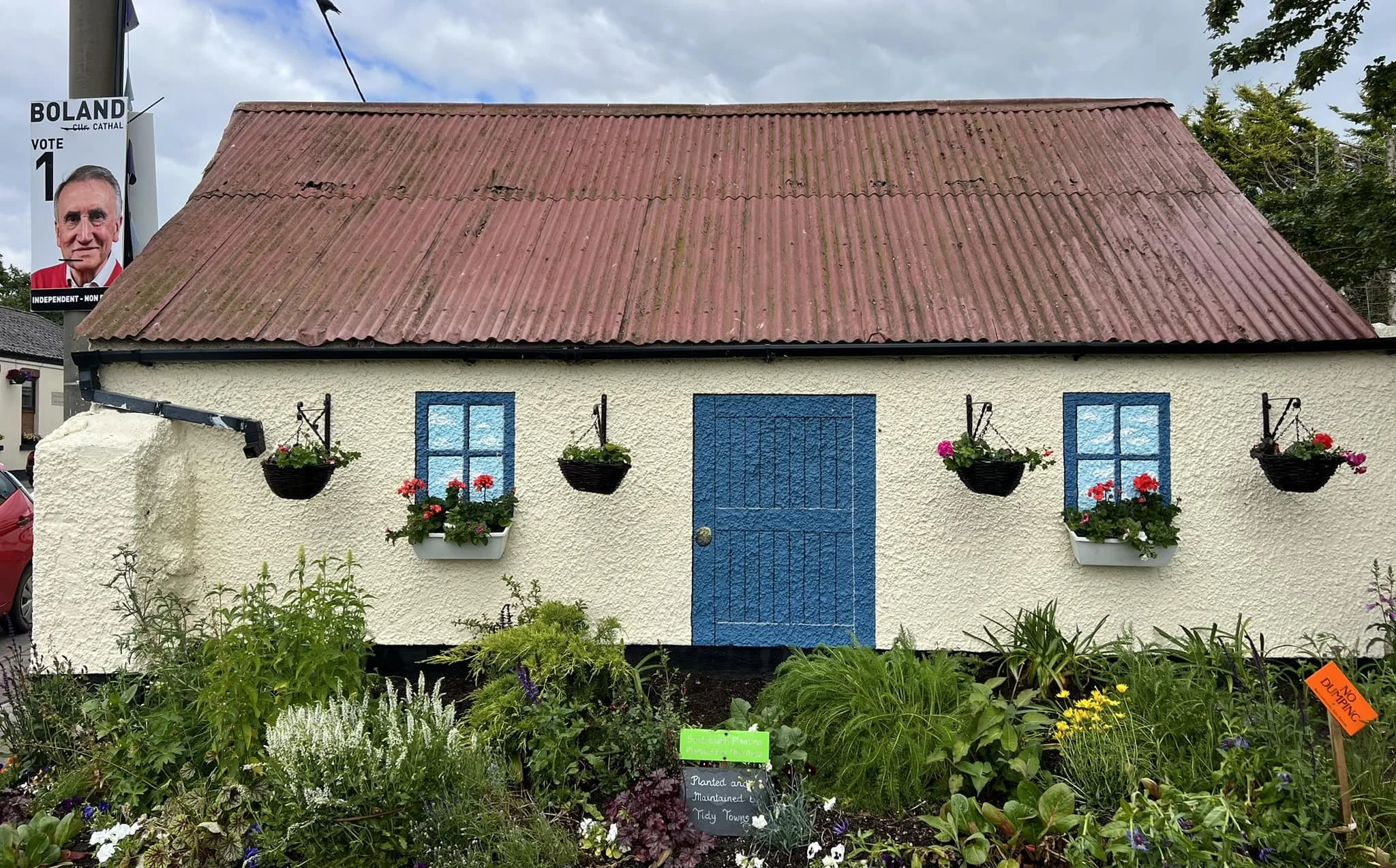 A small, white, textured house with a red corrugated roof and blue window and door frames, surrounded by colorful garden plants and flowers, with hanging black flower baskets on the exterior wall.
