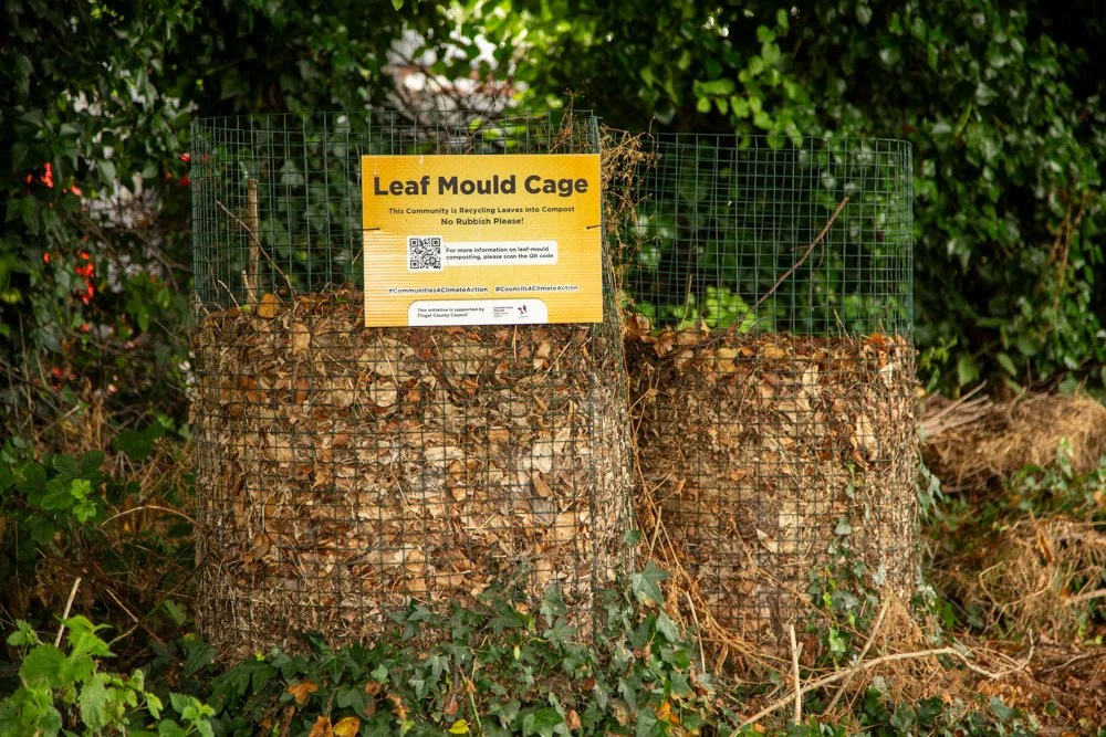 Two tree stumps enclosed by wire cages, each labeled 'Leaf Mould Cage,' with a yellow sign providing instructions for leaf composting, surrounded by green bushes and ivy.
