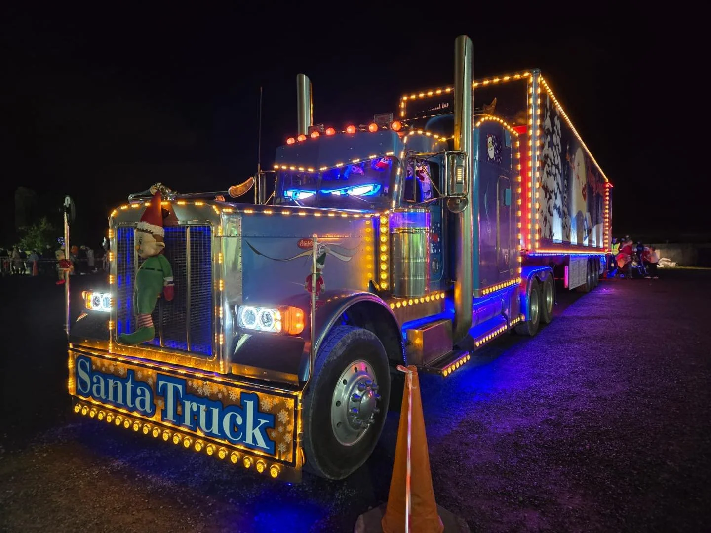 A brightly decorated Christmas-themed truck illuminated with colorful lights at night, featuring a sign that reads 'Santa Truck' and festive decorations including a plush elf figure and Christmas motifs on the side.