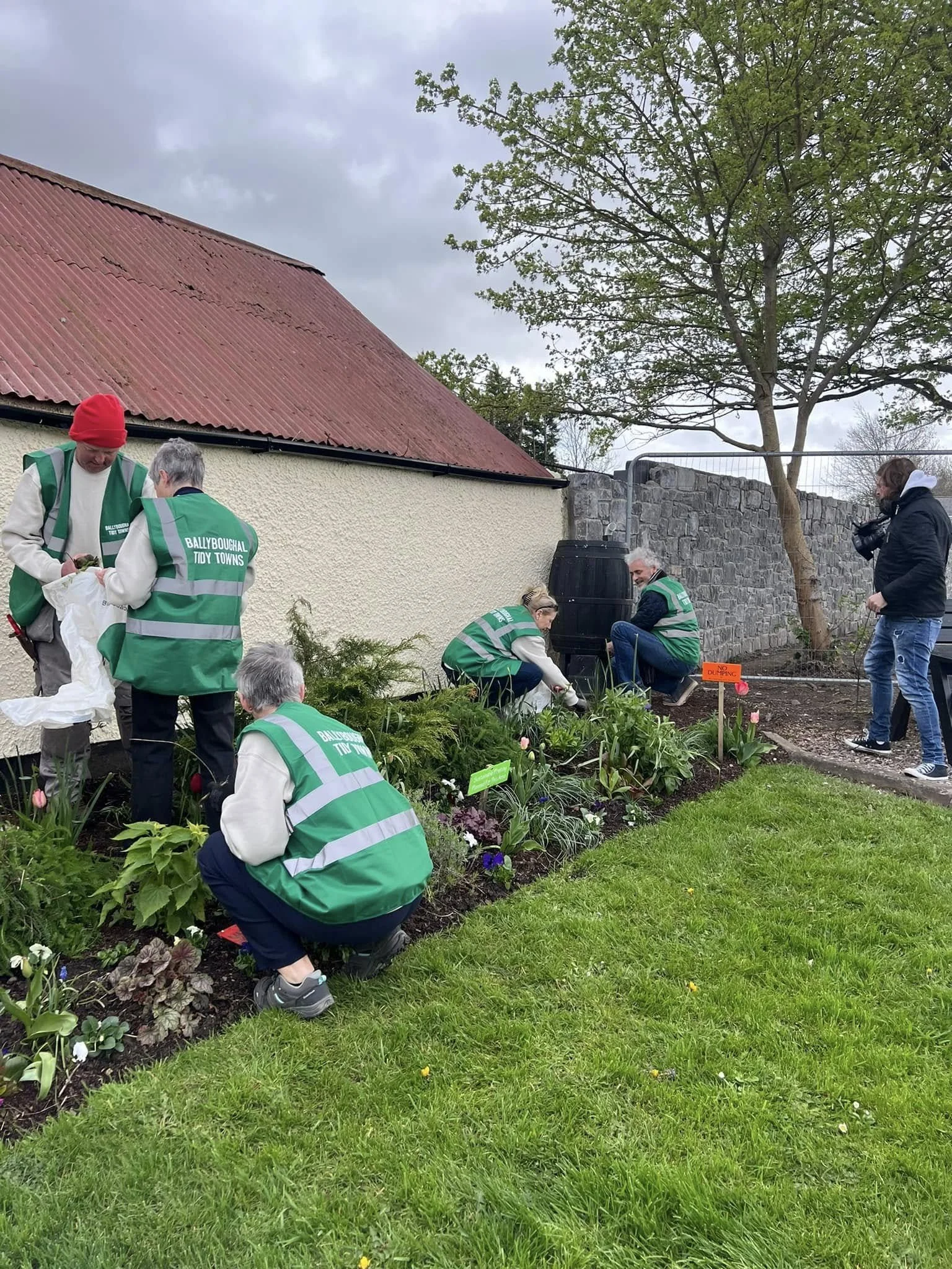 Group of volunteers wearing green vests planting flowers in a garden beside a building with a red roof and a stone wall, while a photographer captures the activity.