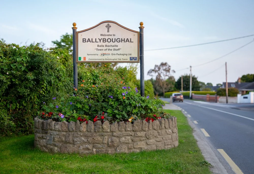 A sign welcoming visitors to Ballyboughal, Ireland, with a flower bed and a roadside in the background.
