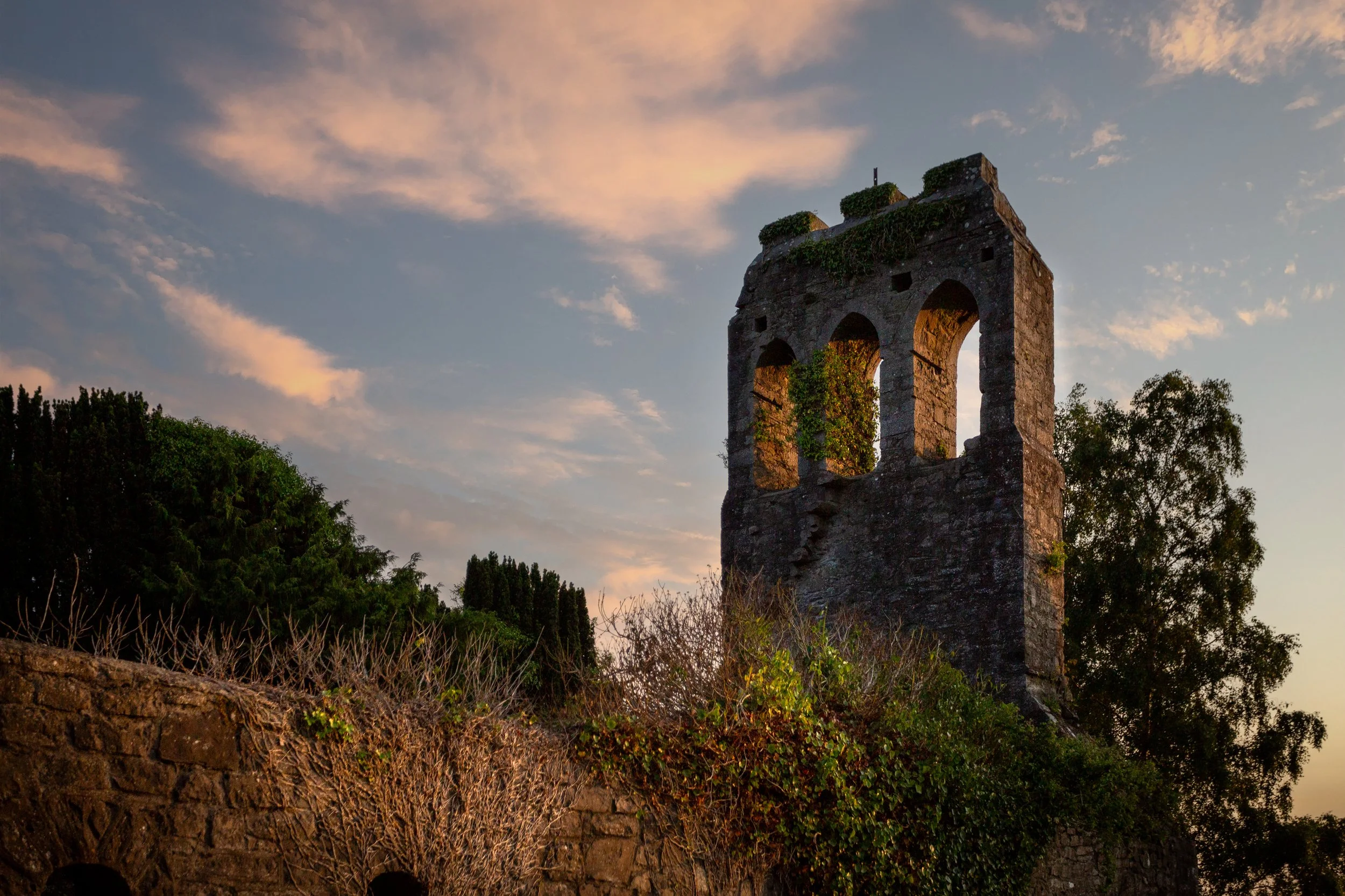 Ruins of an ancient stone tower with three arched windows, ivy growing on it, set against a sunset sky with scattered clouds and trees in the background.