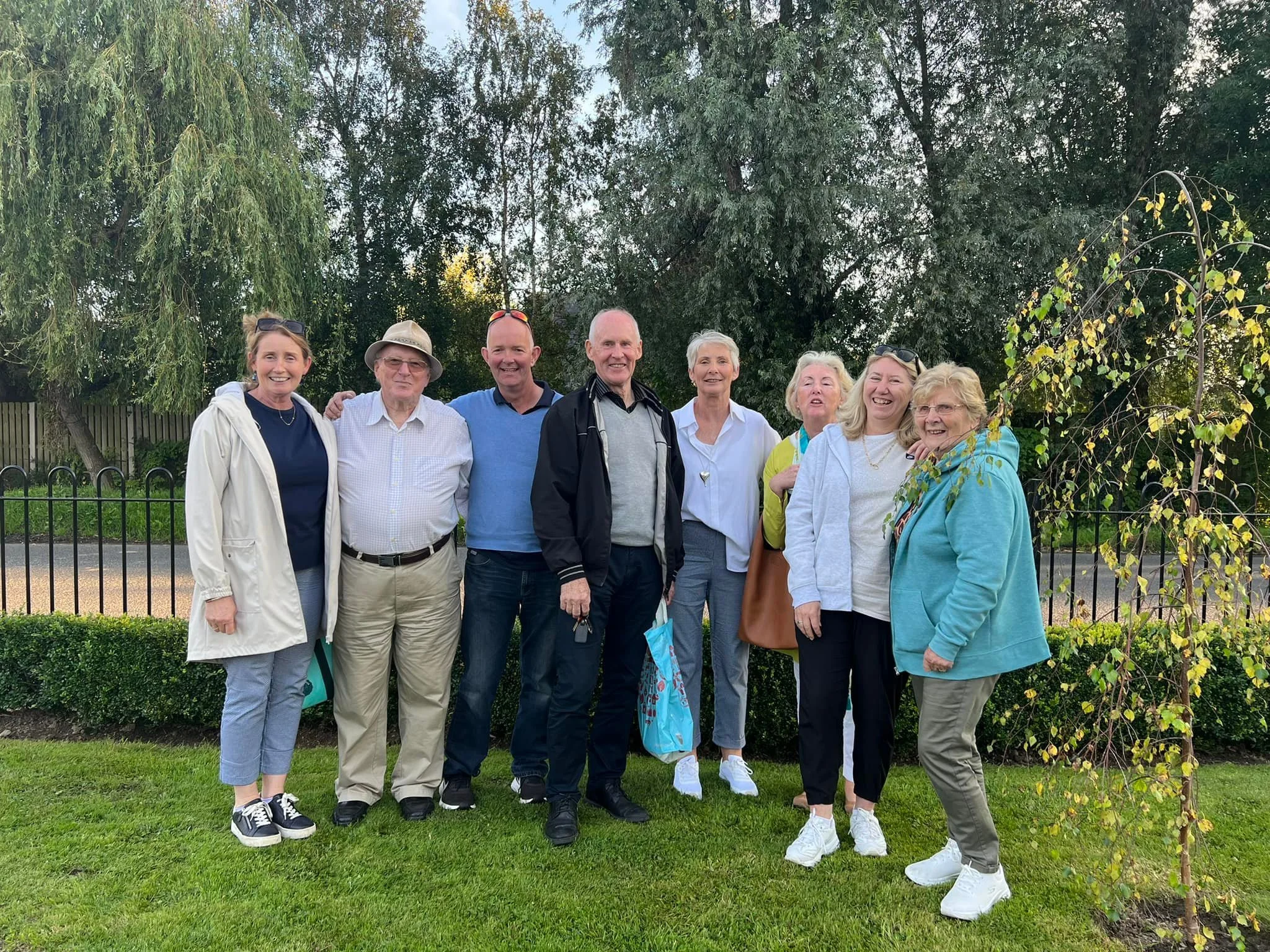 A group of eight older adults, five women and three men, standing outdoors on a grassy area with trees and a fence in the background. They are smiling and posing for the photo.