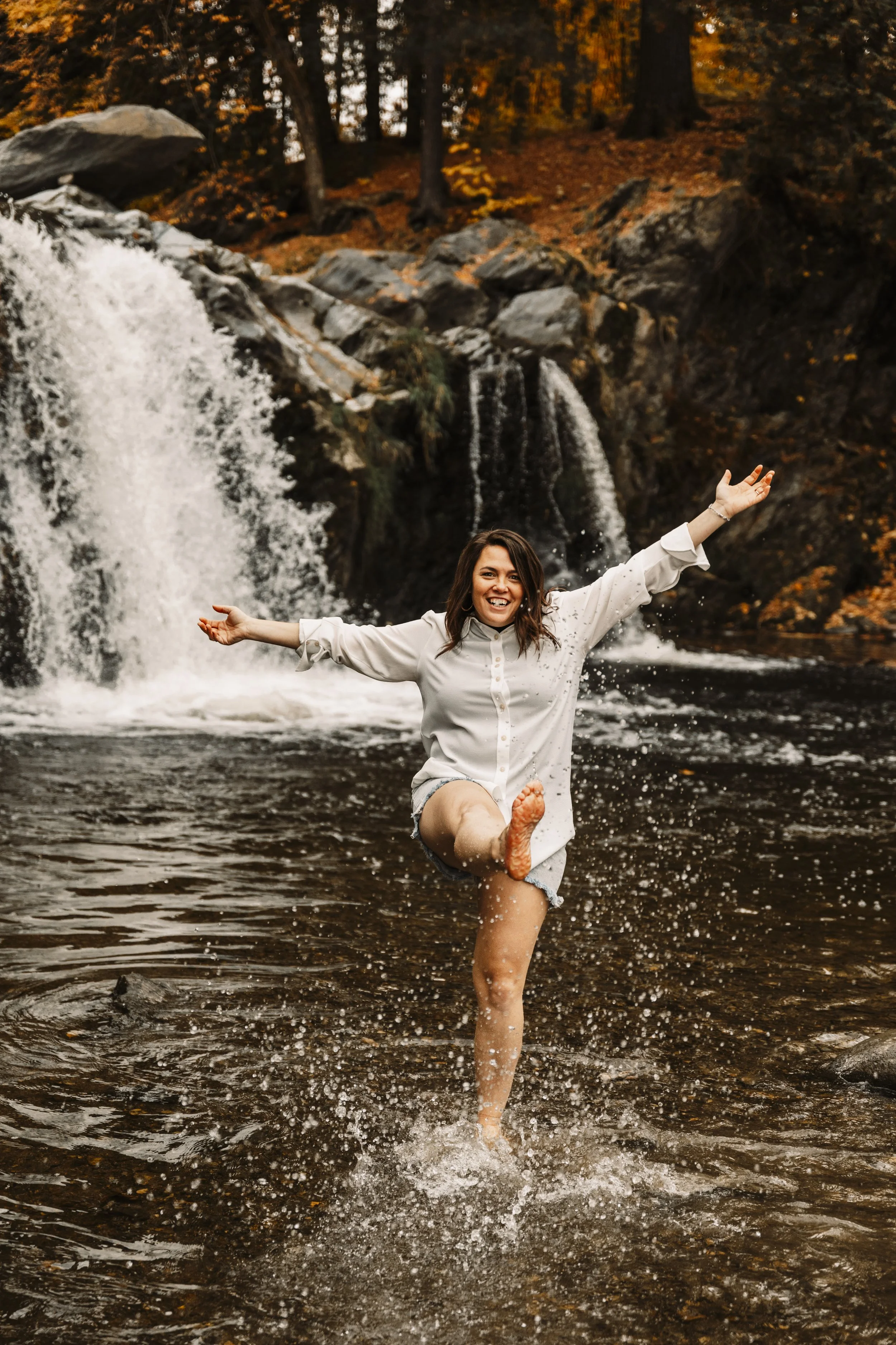 une femme souriante aux cheveux longs et bruns, portant un haut blanc et se tenant devant une chute d'eau exprimant la joie et la liberté"
