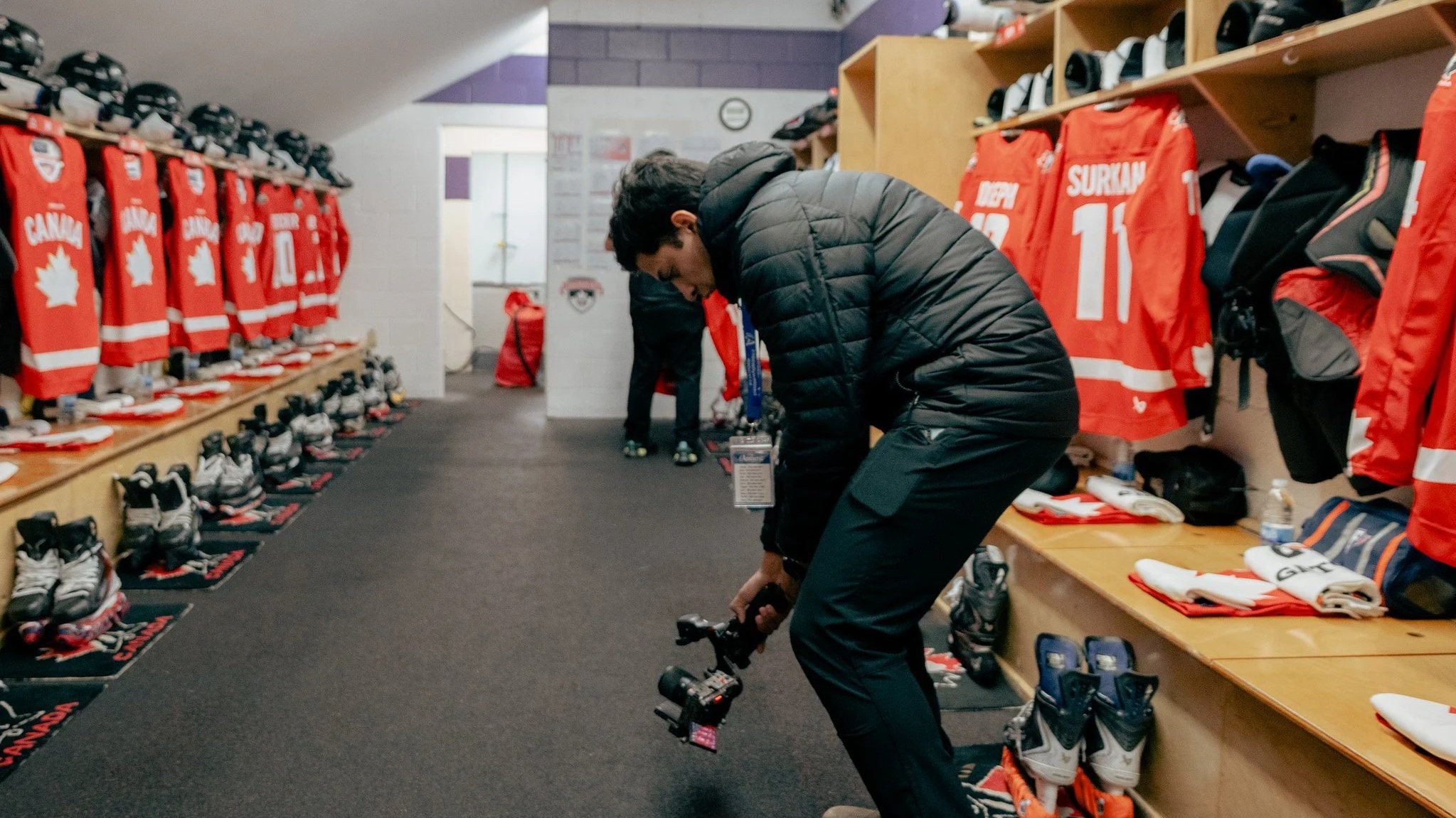 A hockey locker room with Canadian hockey jerseys, helmets, skates, and equipment. A man in a black jacket is adjusting a camera, bending down in the middle of the room.