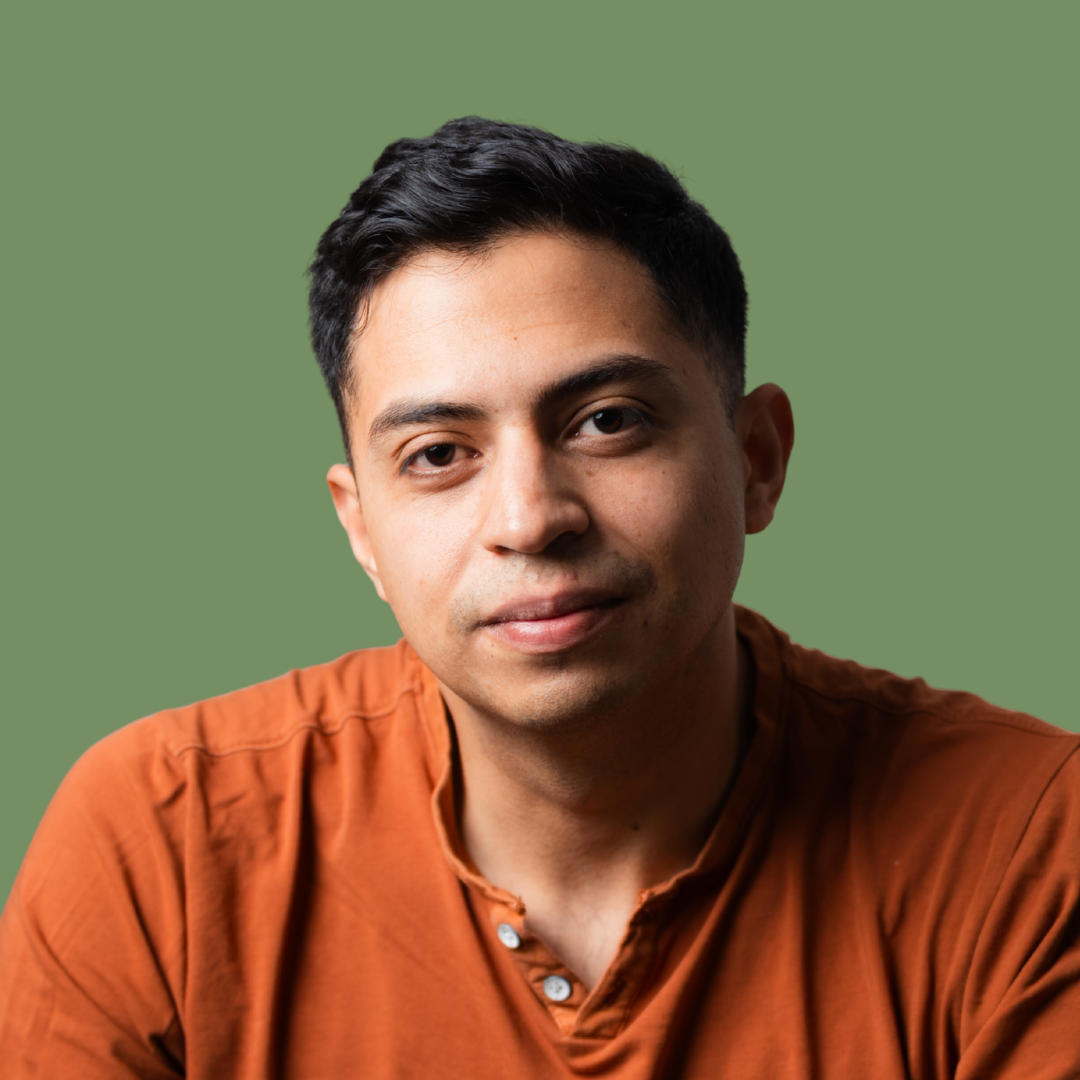 A young man with short black hair and light skin, wearing an orange shirt, posing against a plain green background.