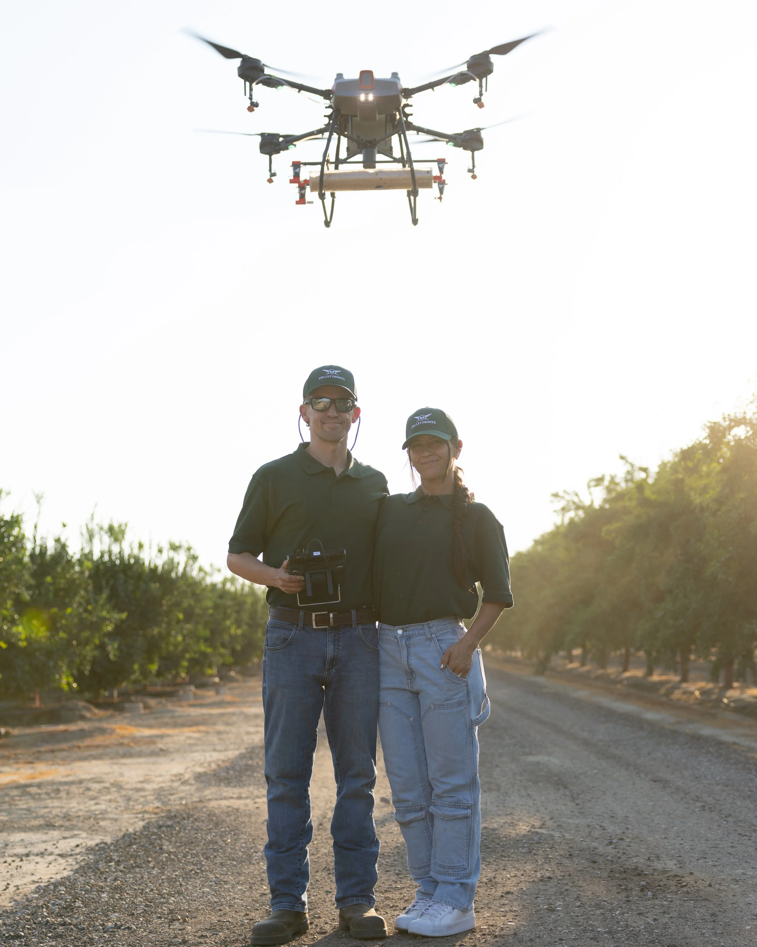 Two drone operators standing on a dirt road holding a remote control, with a drone flying overhead in sunny outdoor setting.