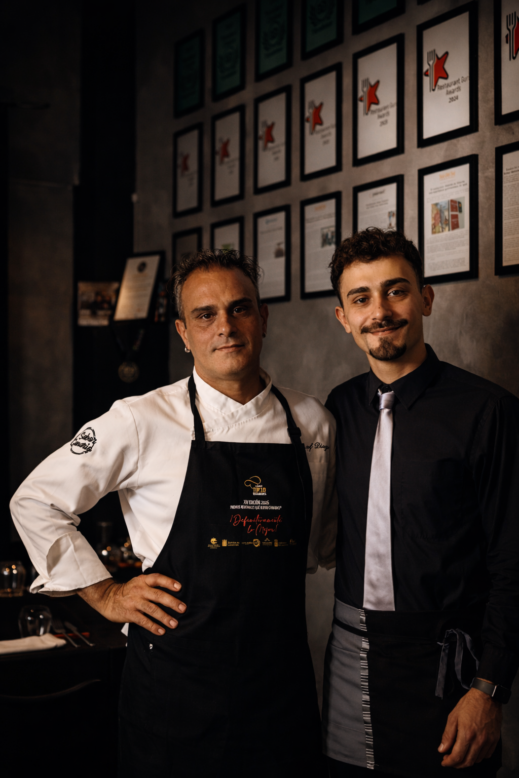 Dos hombres, uno chef y otro posiblemente camarero, posando juntos en un restaurante con paredes decoradas con certificados y premios.
