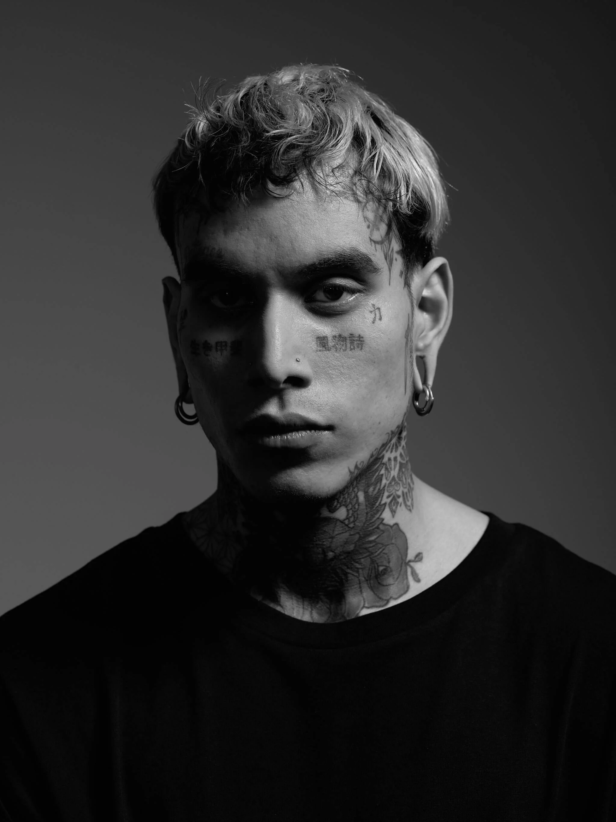 Retrato en blanco y negro de un joven con pelo rubio y rizado, con tatuajes en el rostro y cuello, usando pendientes grandes en las orejas, contra un fondo oscuro.
