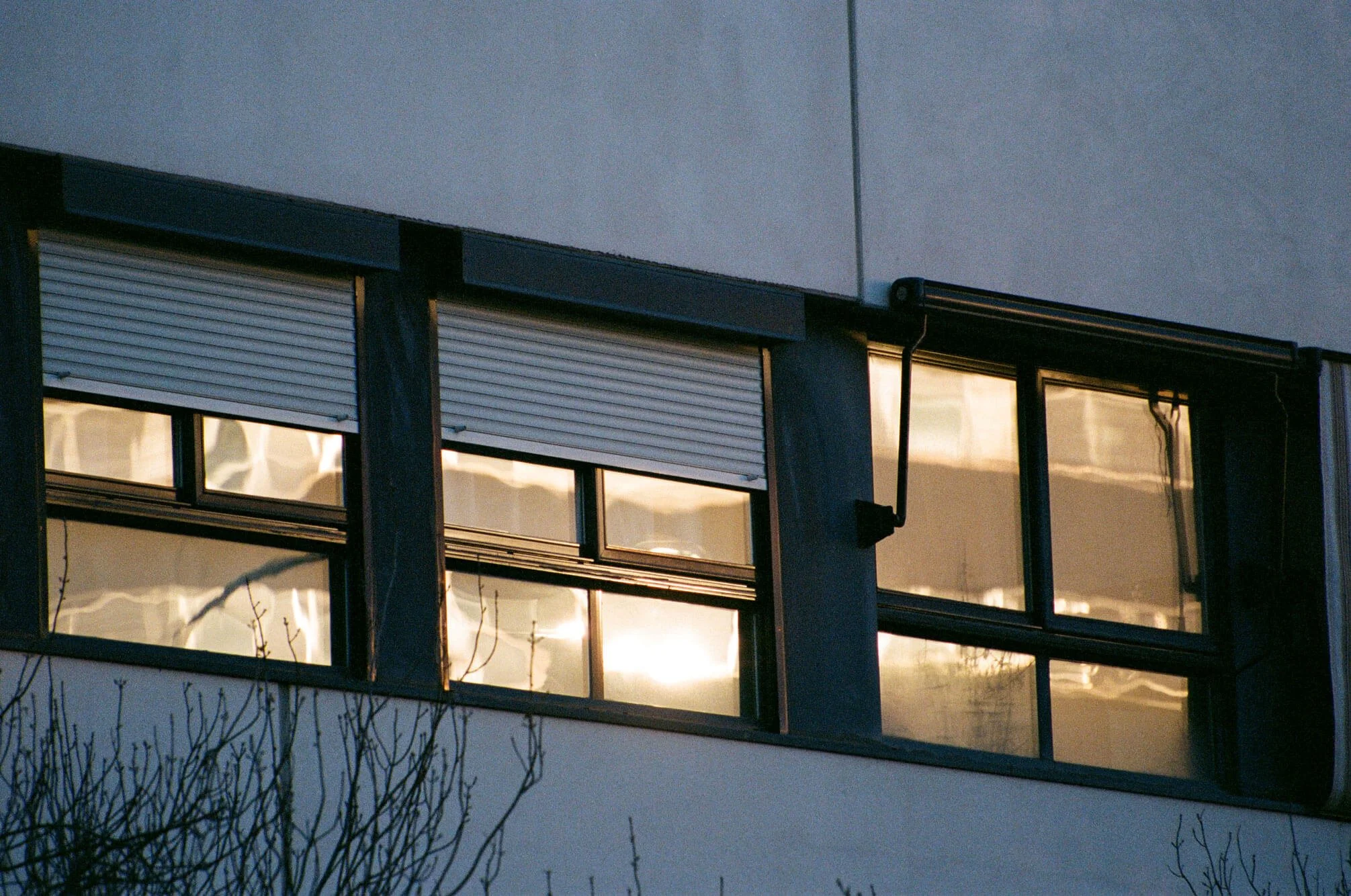 Vista de una fachada de edificio con ventanas y persianas. La iluminación interior resalta a través de las ventanas, y en la parte superior hay una canaleta de desagüe. Al frente, hay ramas de árboles sin hojas.