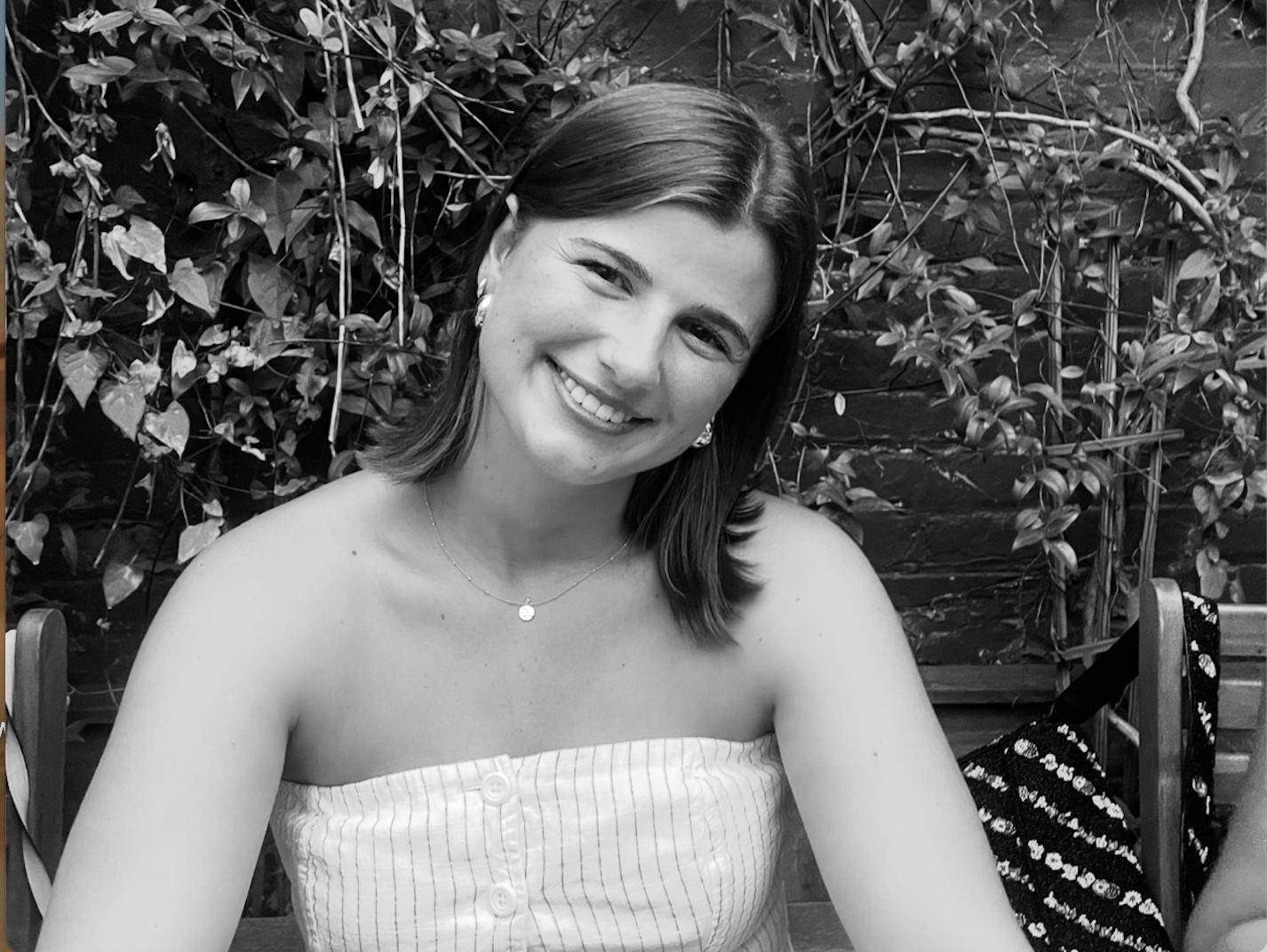 A young woman smiling, sitting outdoors with a background of leafy vines.