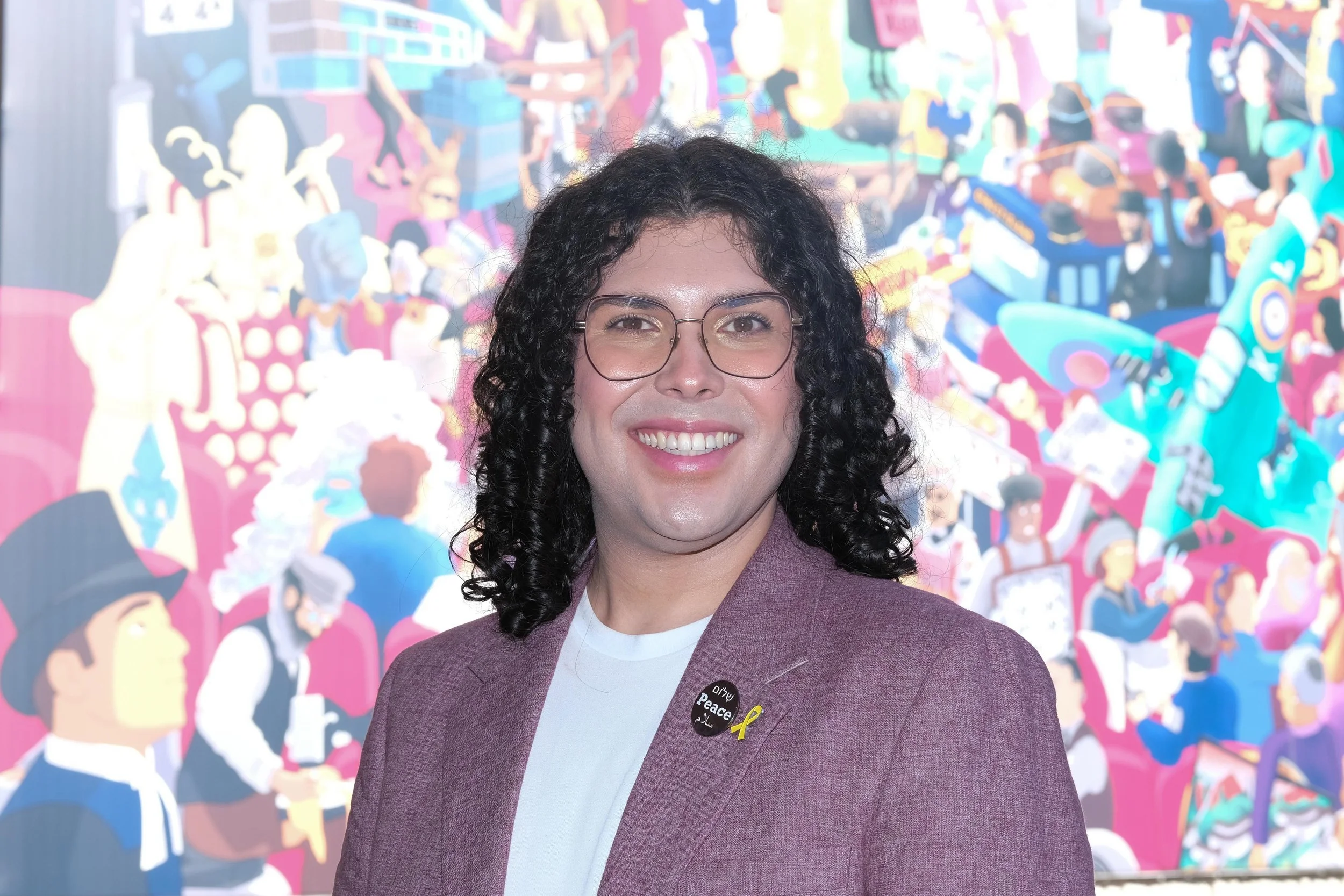 A smiling person with long curly dark hair, wearing glasses, a burgundy blazer, and a white shirt, standing in front of a colorful mural.
