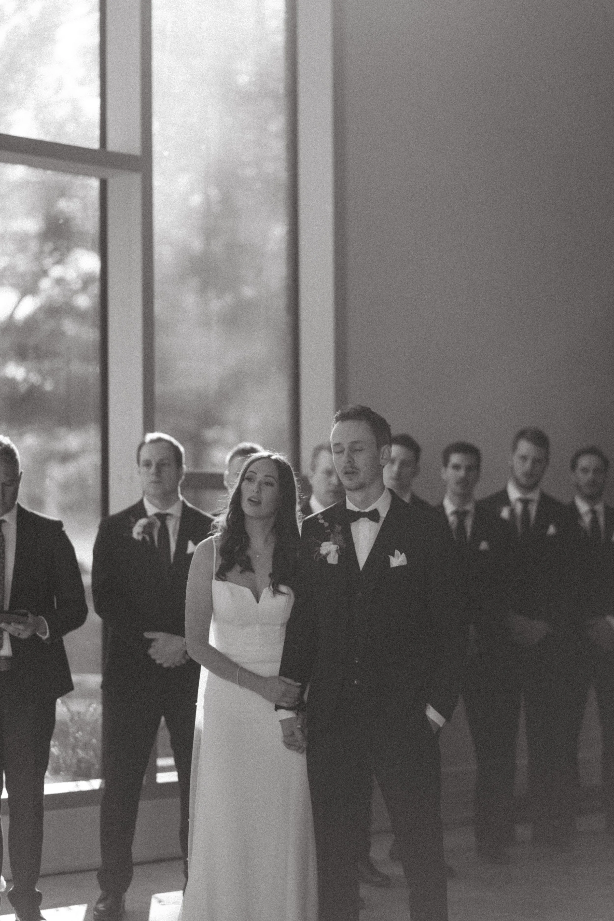 Black and white photo of a wedding ceremony, with a bride and groom holding hands, surrounded by groomsmen and bridesmaids, in a room with large windows.