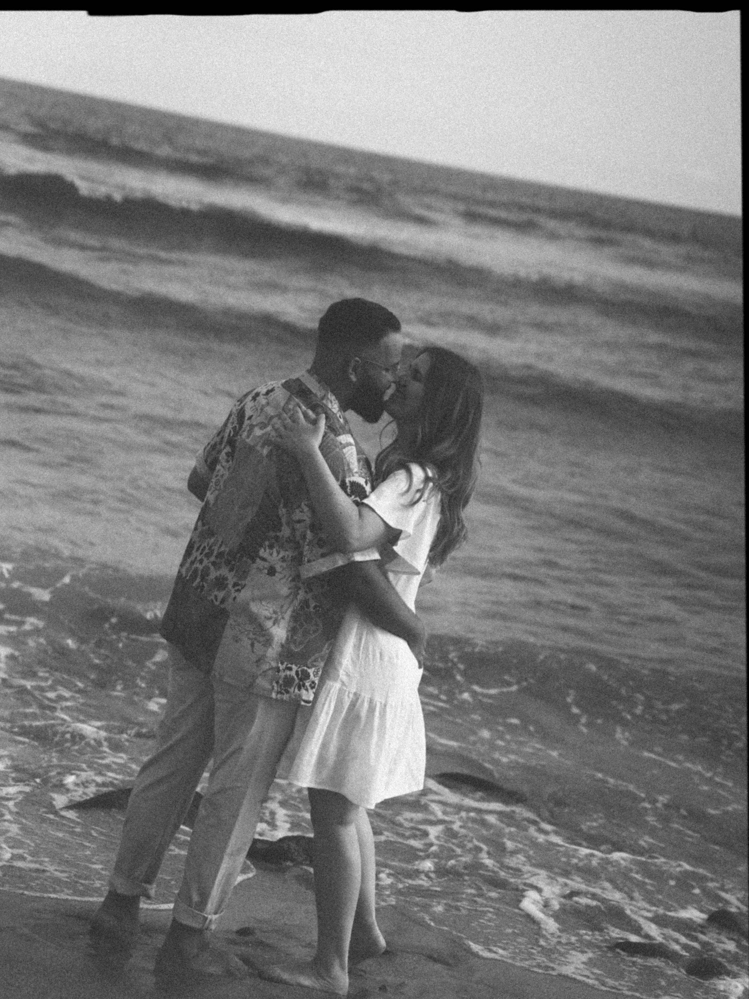 A black and white photo of a couple embracing on the beach, near the ocean waves.