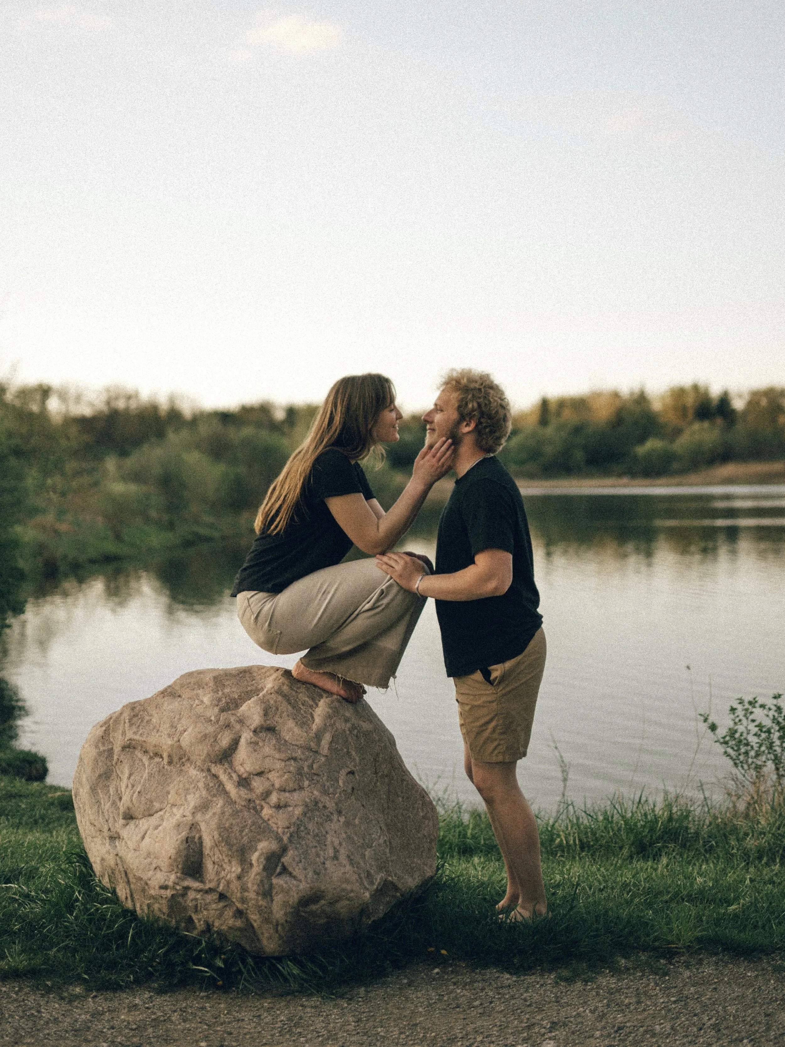 A couple by a lake, the woman sitting on a large rock with her legs around the man's shoulders, smiling and gazing at each other.