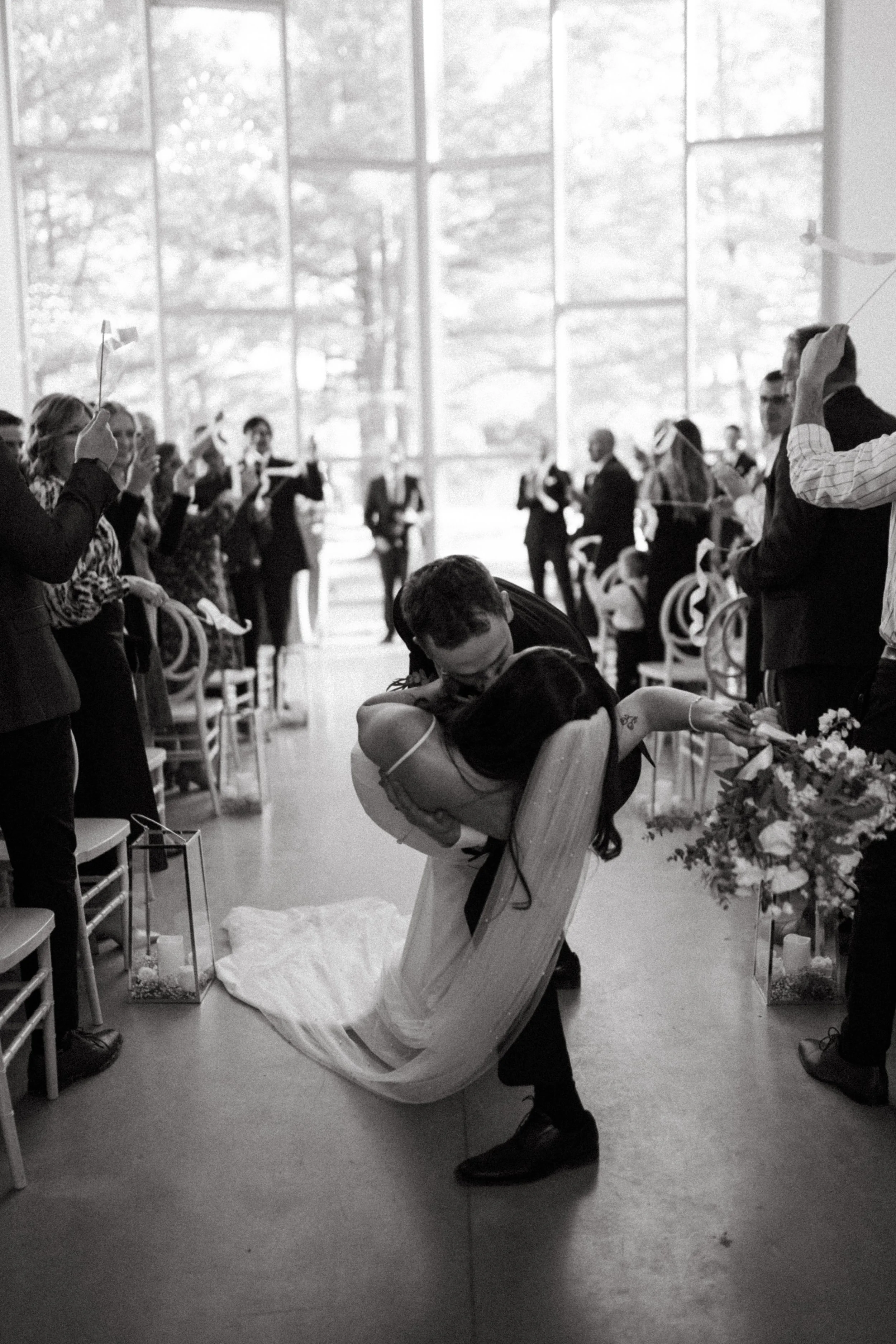 A couple dancing closely at their wedding reception, surrounded by guests holding small flags and celebrating in a bright, modern venue.