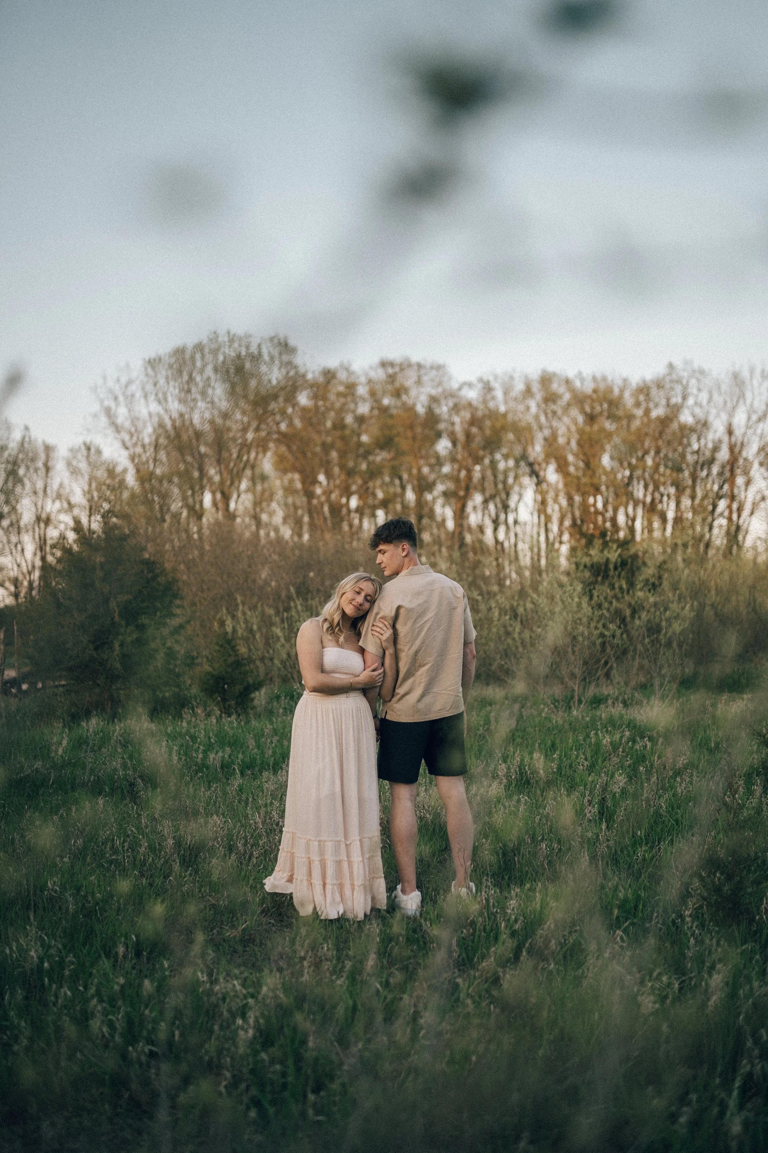 A young couple standing in a grassy field during sunset, with the woman hugging the man and both smiling softly, surrounded by trees in the background.