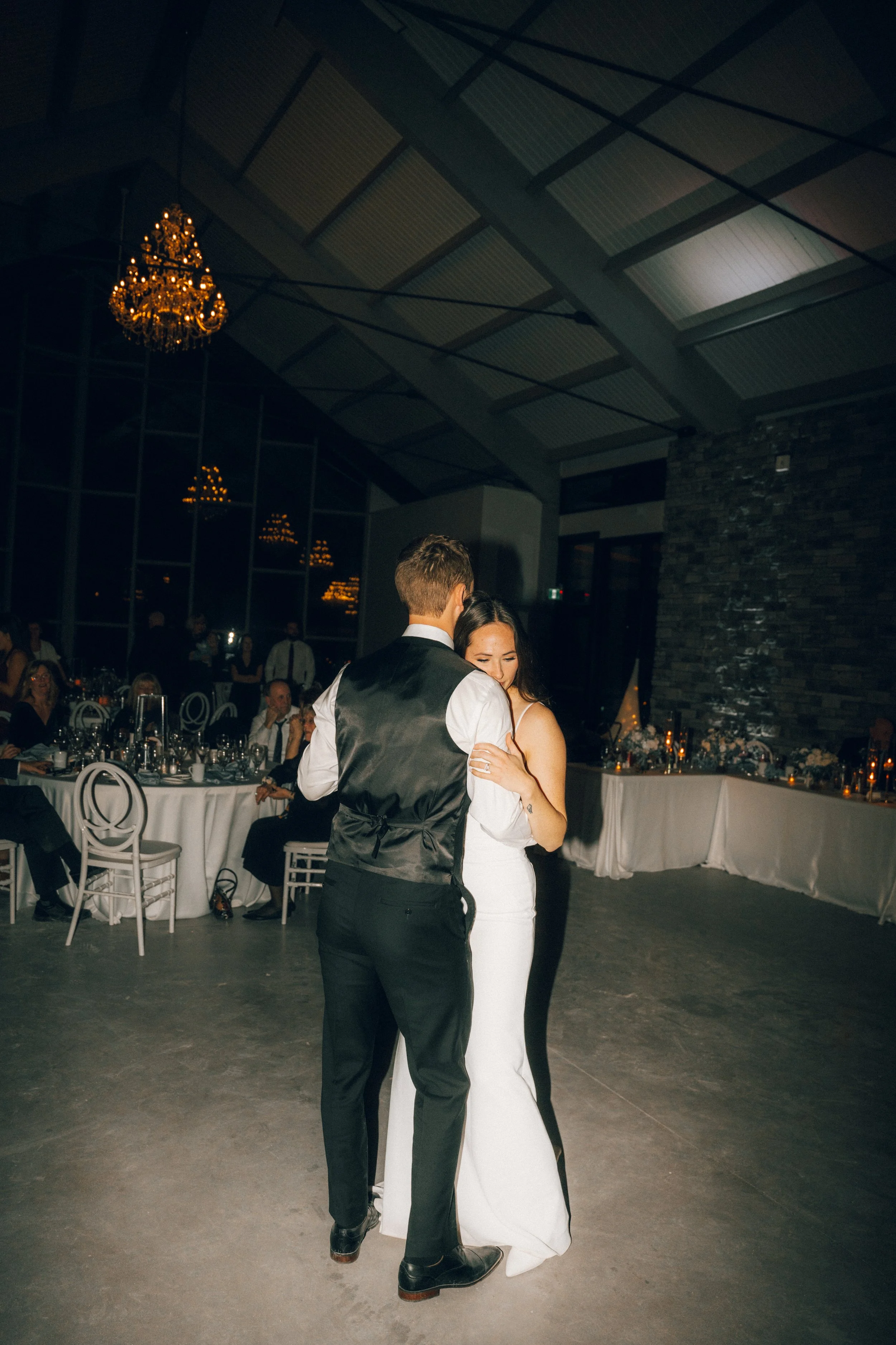 A bride and groom are dancing closely at their wedding reception in a dimly lit hall with elegant decor and seated guests in the background.
