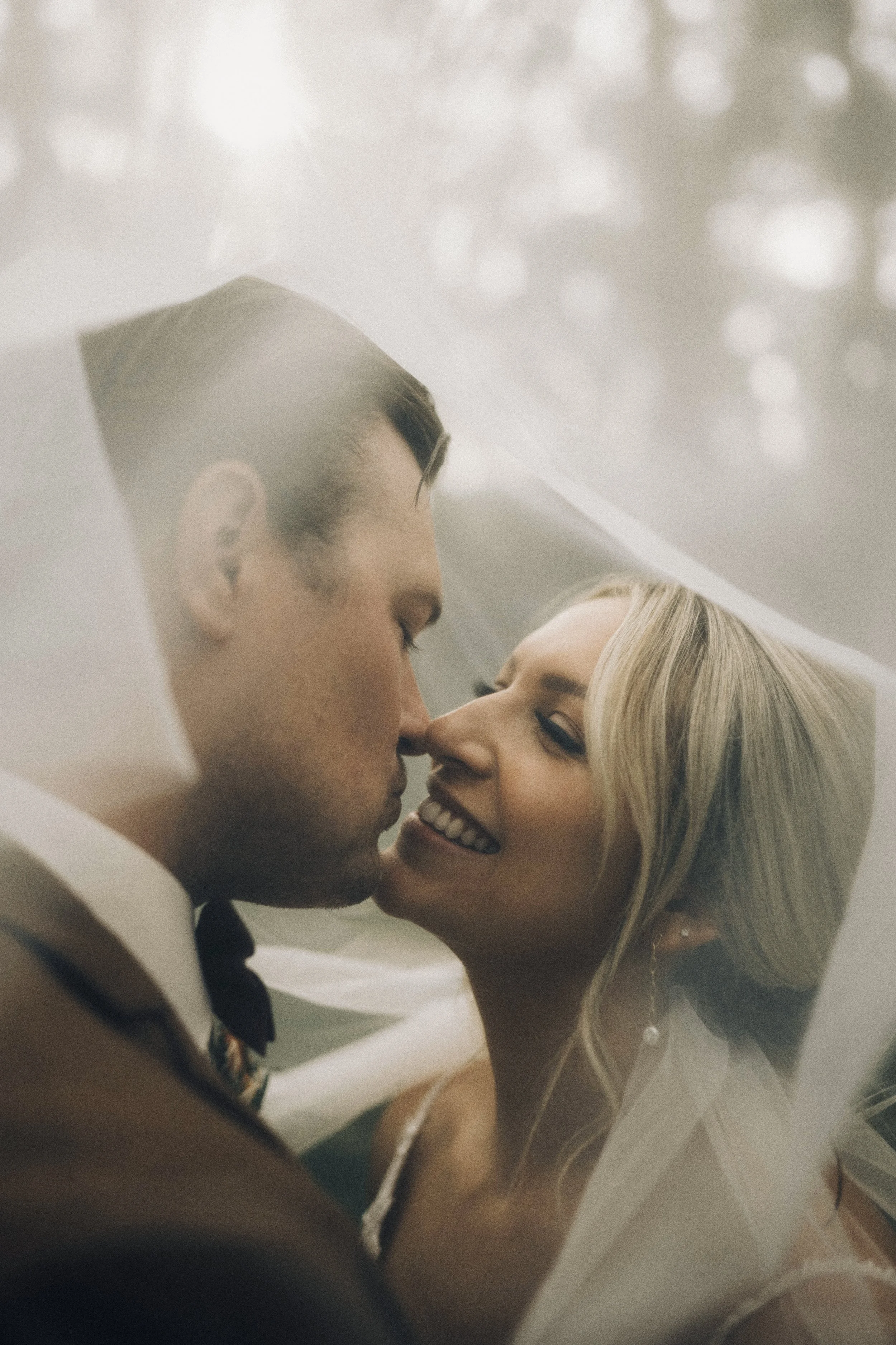 A bride and groom sharing a kiss under a veil, with soft lighting and the bride smiling in Chatham, Ontario