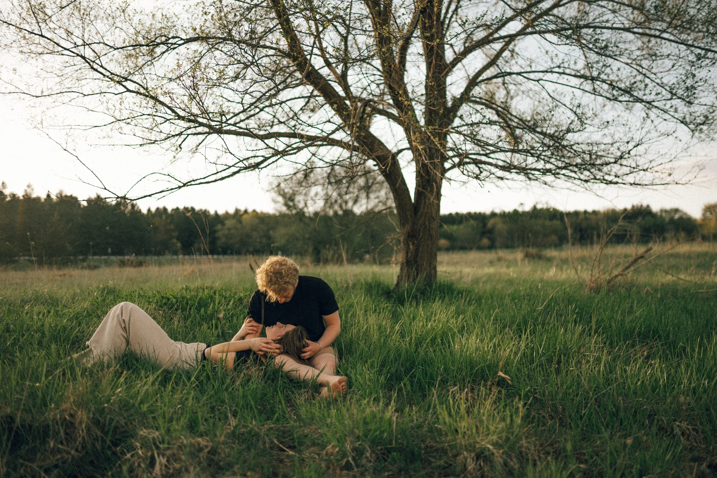 A man and woman lying on the grass under a leafless tree in a field, with the man leaning over the woman, appearing to comfort her.