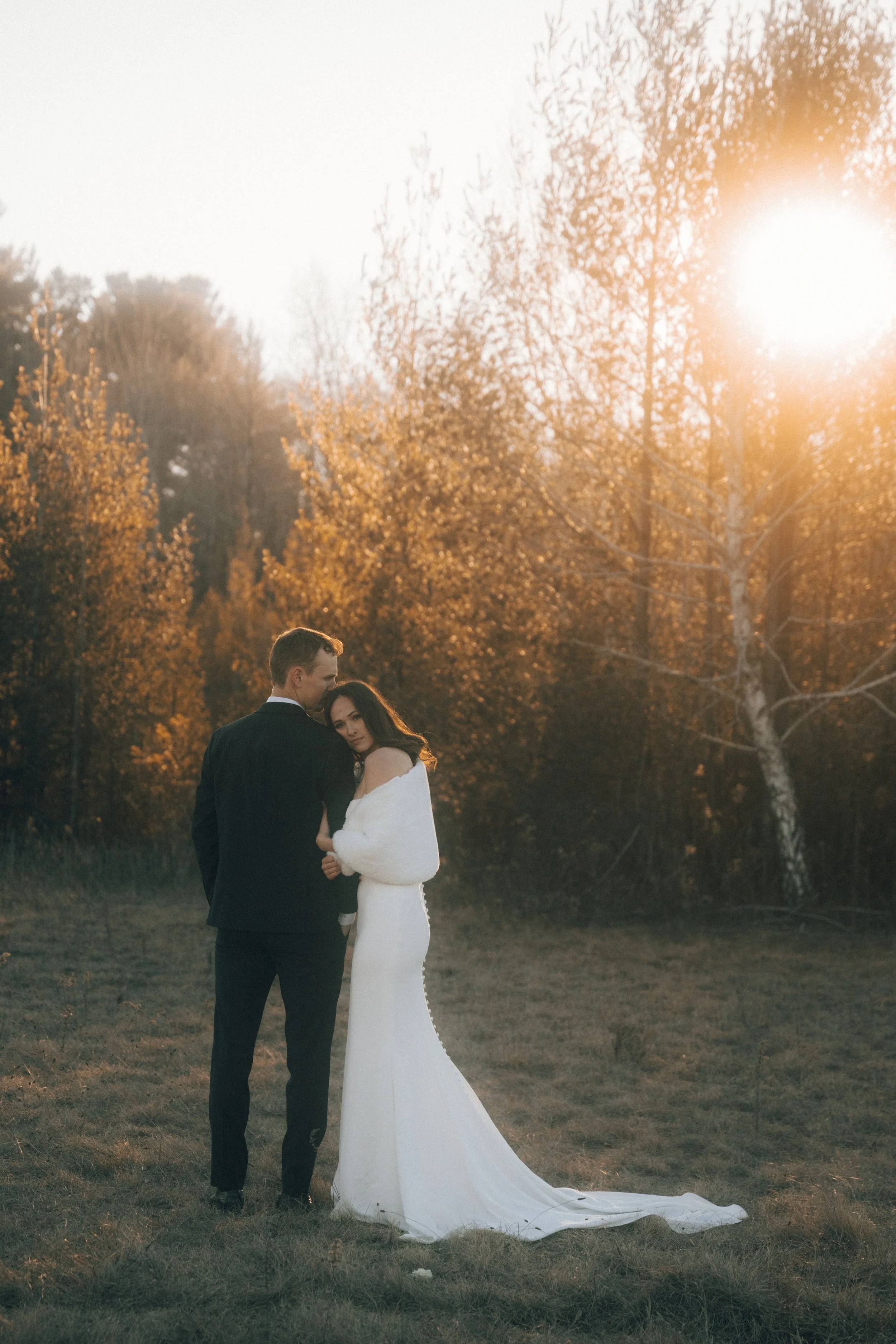 A bride and groom standing in a grassy field during sunset, with trees in the background and the sun shining brightly behind them.