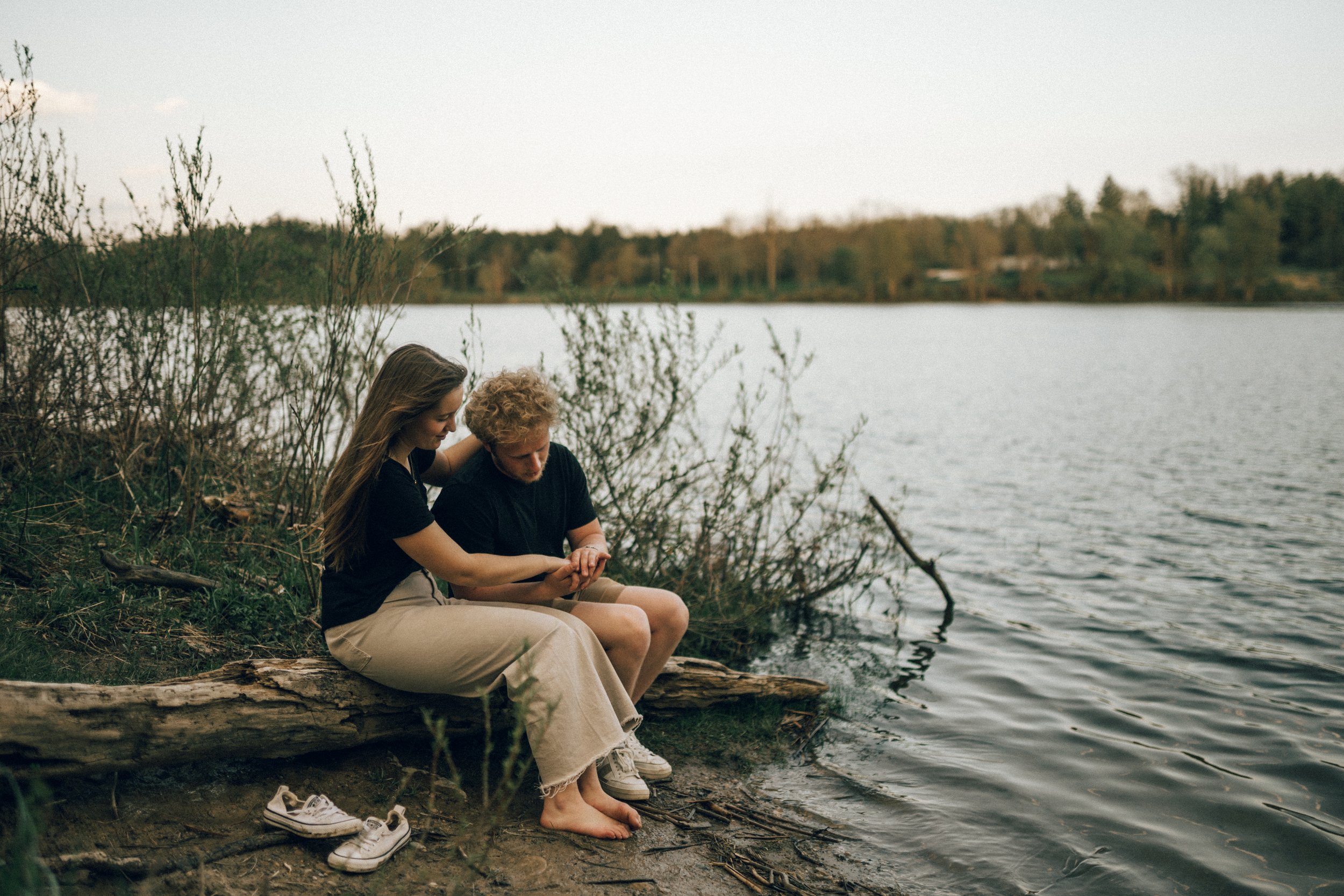 A young couple sitting by a lakeshore, looking at a phone together, with shoes on the ground nearby, surrounded by bushes and trees under a clear sky.