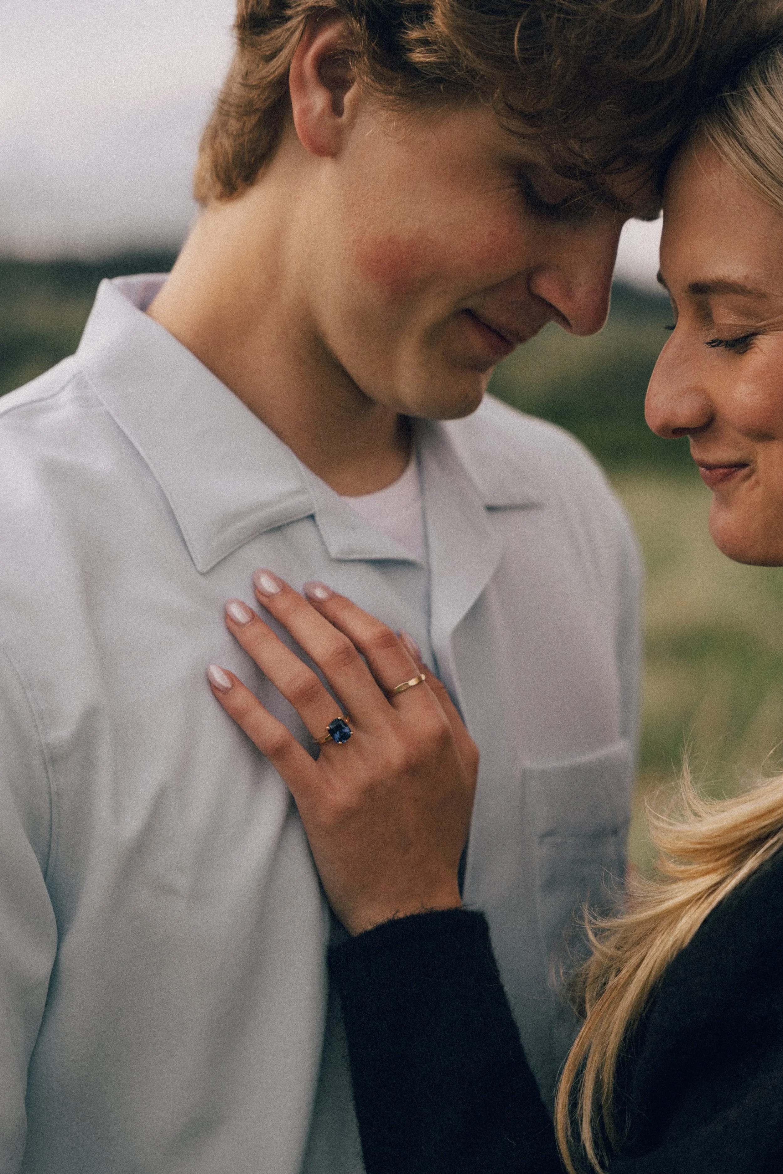 A couple with their foreheads touching, smiling softly, with the woman's hand resting on the man's chest showing an engagement ring with a large blue gemstone.