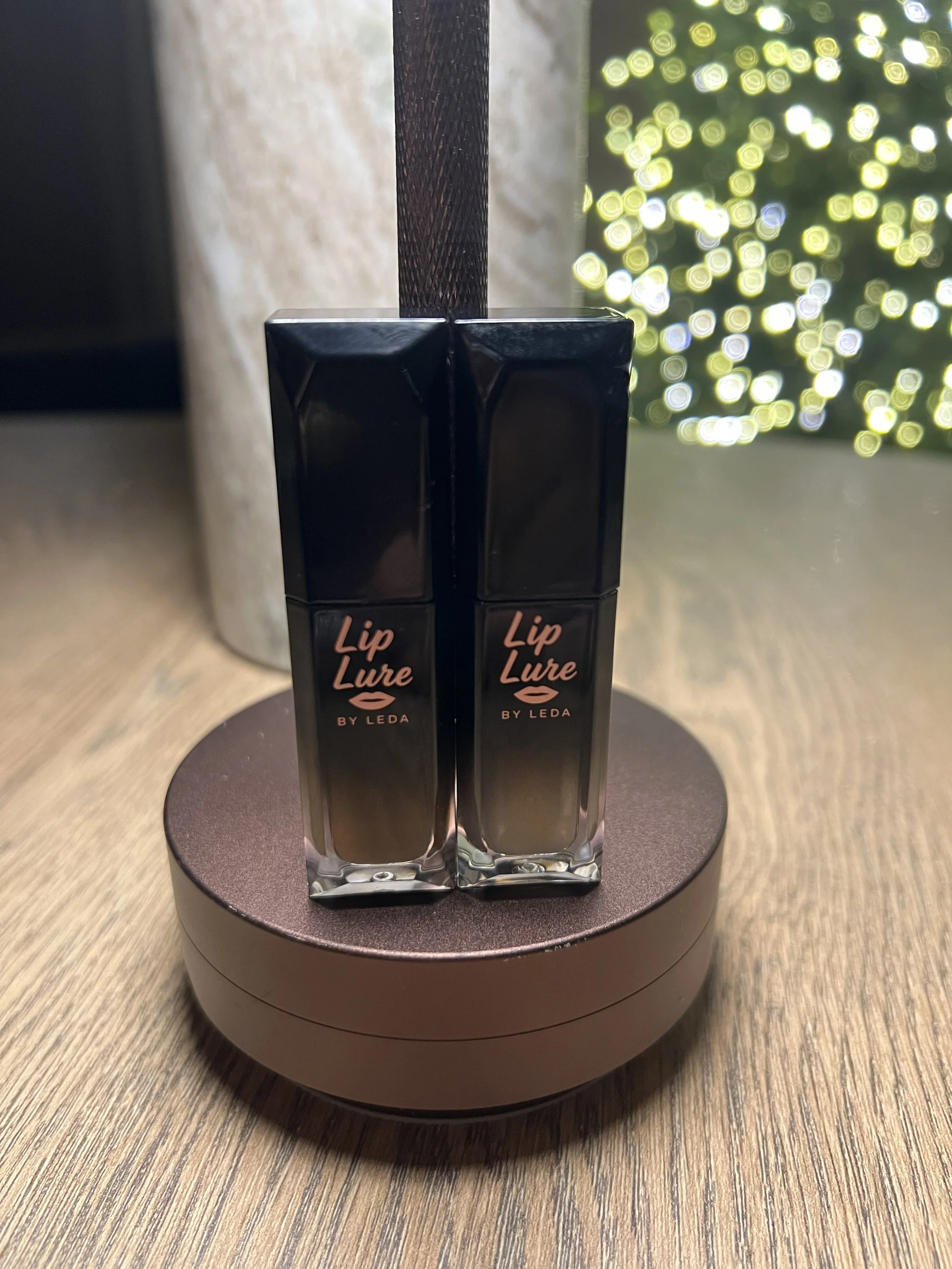 Two lip gloss bottles labeled 'Lip Lure by Leda' standing upright on a round, dark pink makeup case on a wooden surface, with a curtain of bokeh lights in the background.