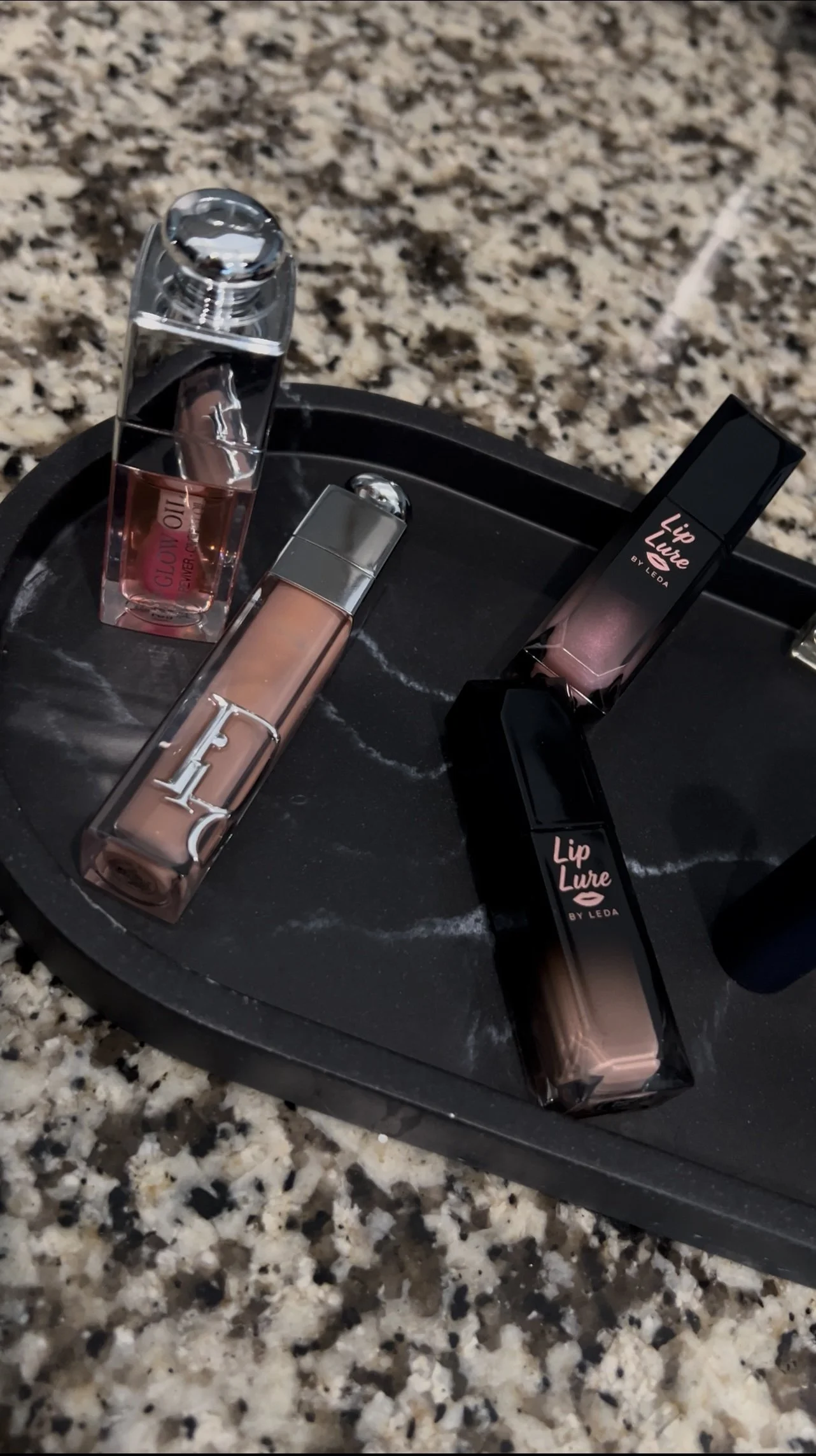 Various lip glosses with black and clear caps on a black tray, placed on a granite countertop.