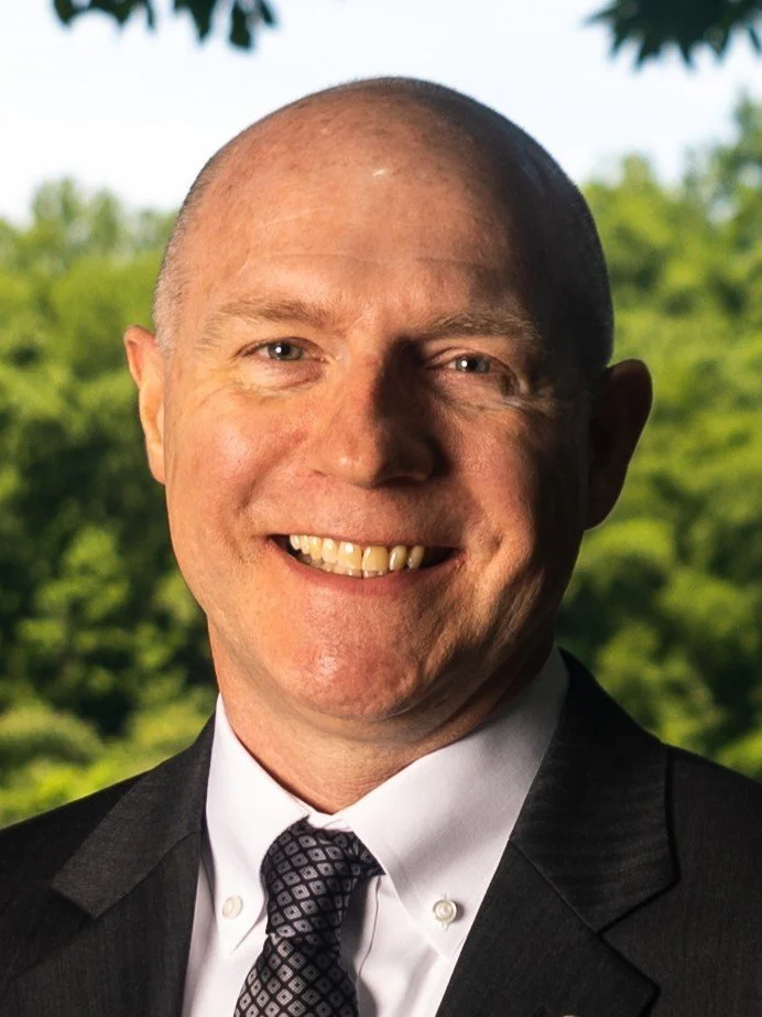 Man in a suit smiling outdoors with greenery in the background.