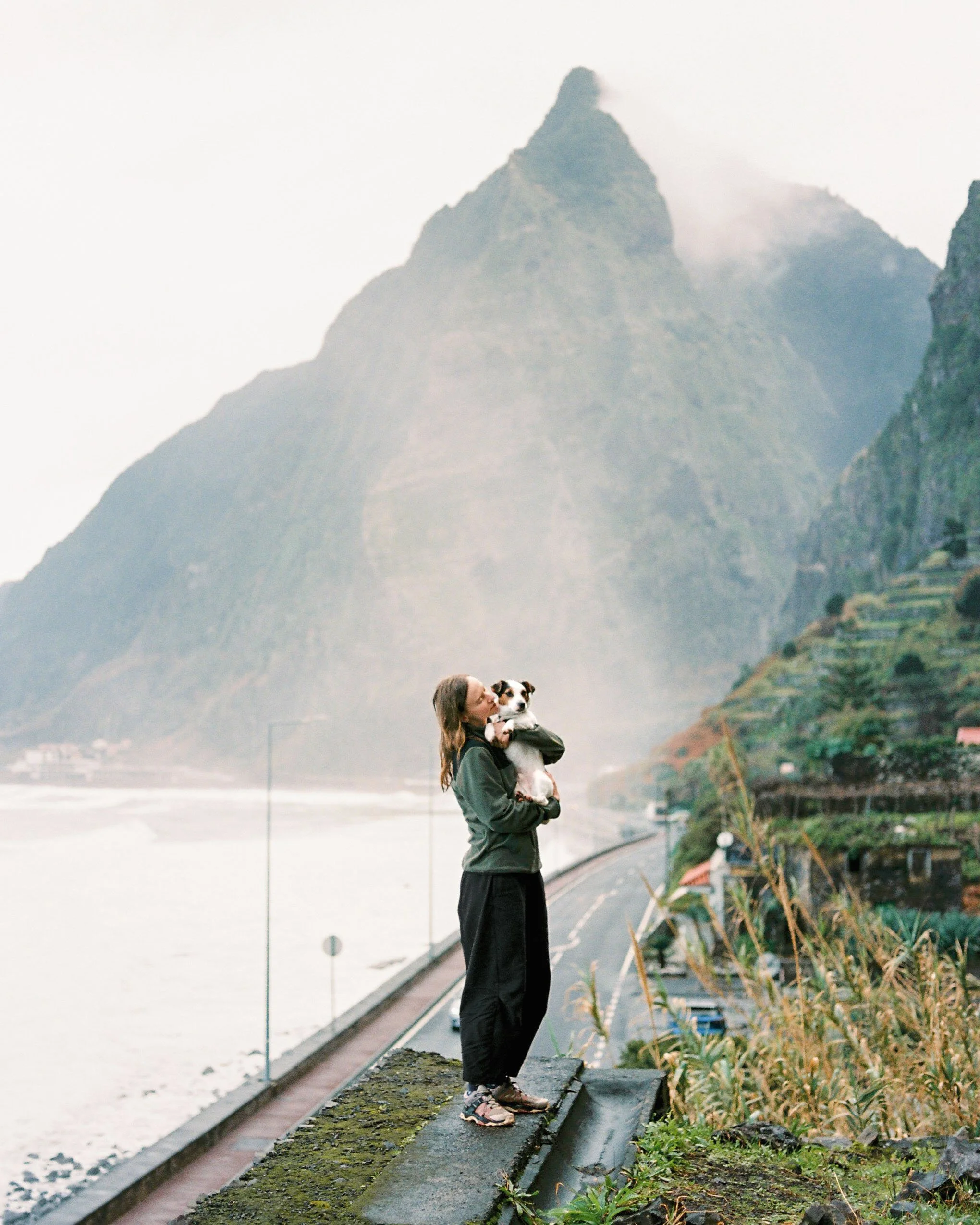 A fine art 35mm film portrait of a Jack Russell Terrier dog and the owner standing in front of green mountain in Madeira and the Atlantic ocean.