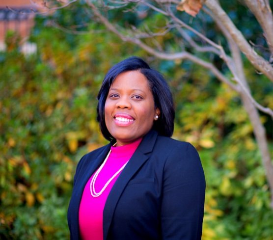 A woman with shoulder-length black hair, wearing a black blazer over a pink top and a pearl necklace, smiling outdoors with green and yellow foliage in the background.