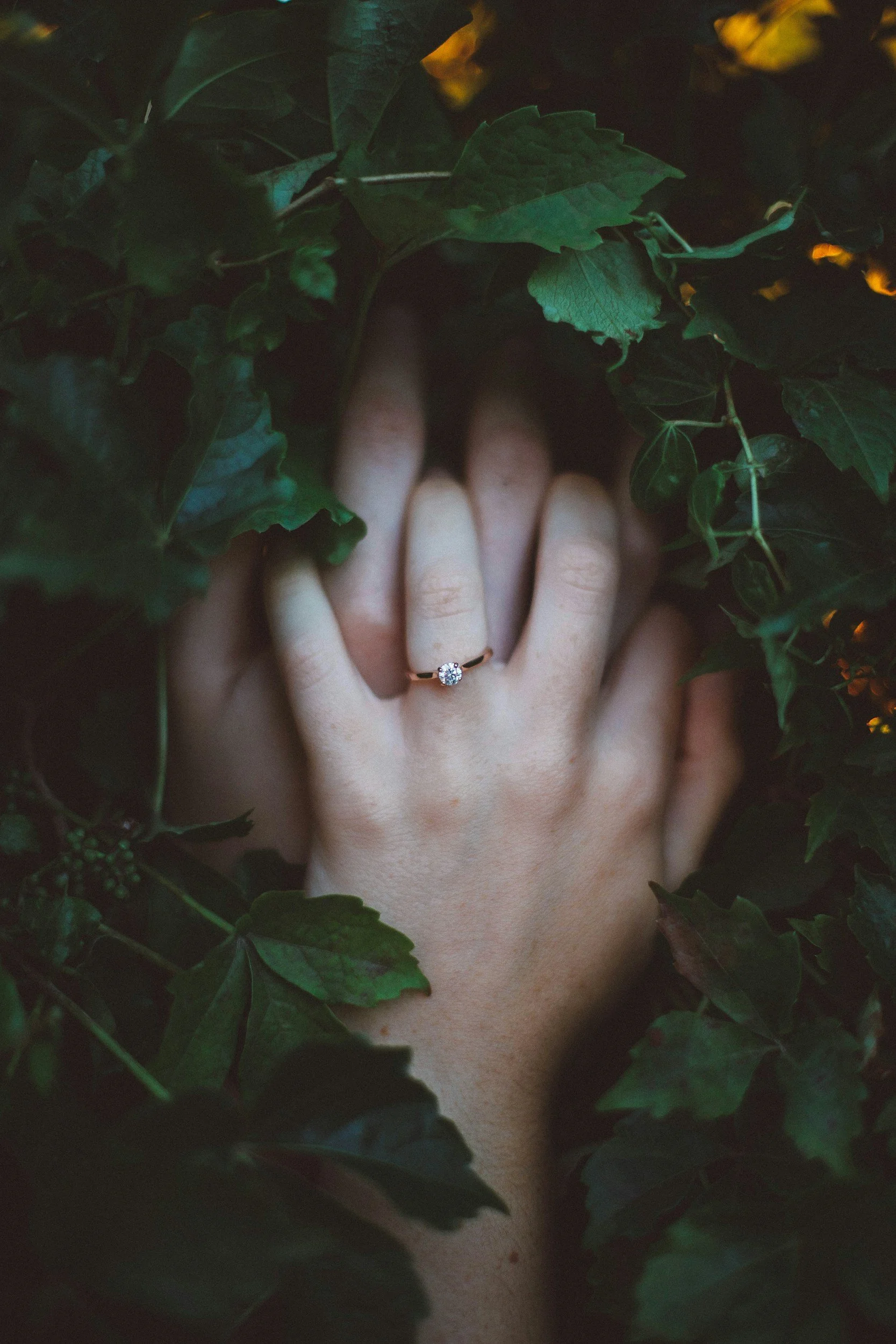 Close-up of a person's hand with an engagement ring, surrounded by green leaves and foliage.