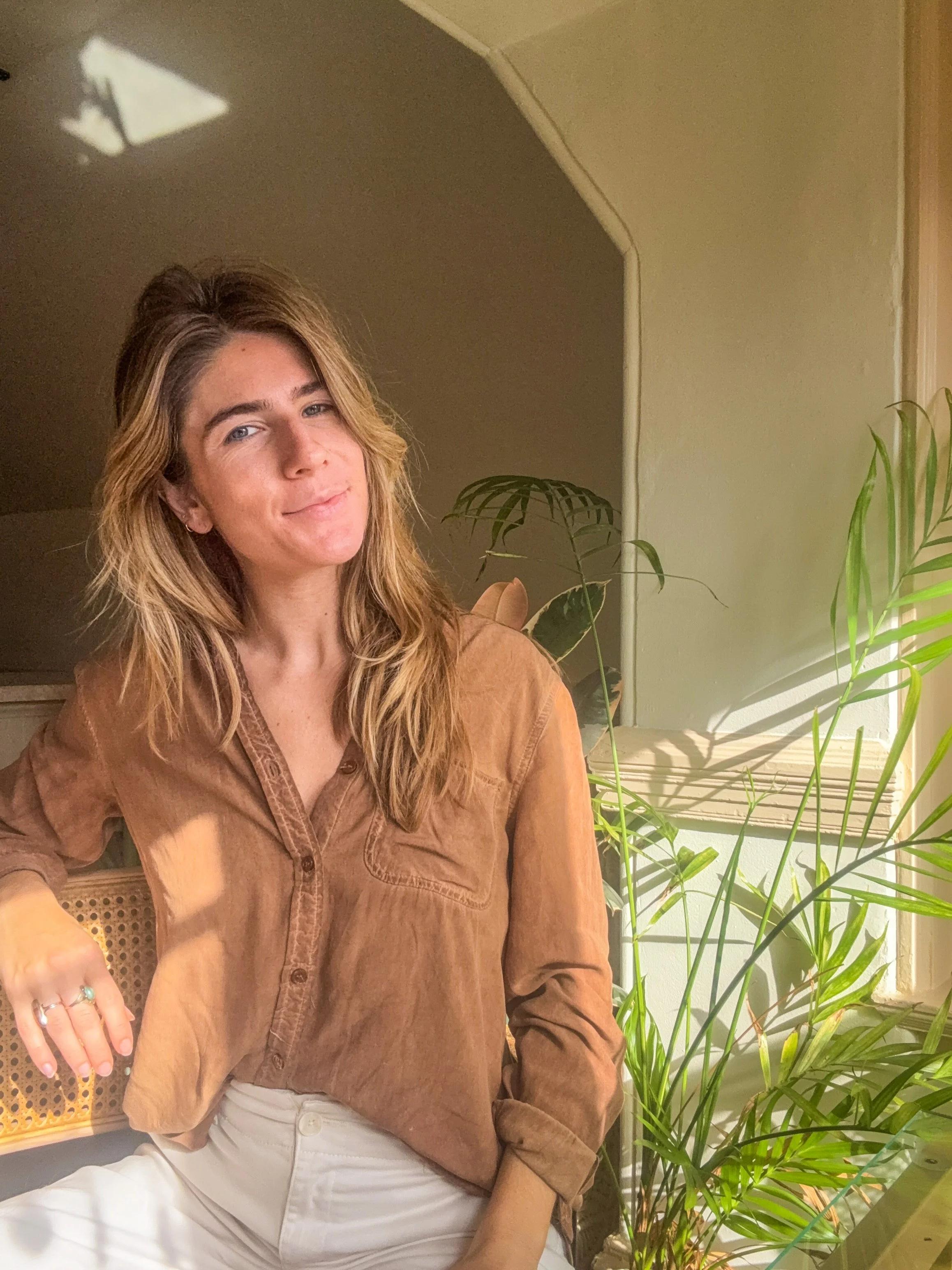 A woman with wavy blonde hair sitting in a room near a window with sunlight, next to green houseplants, wearing a brown button-up shirt and white pants.