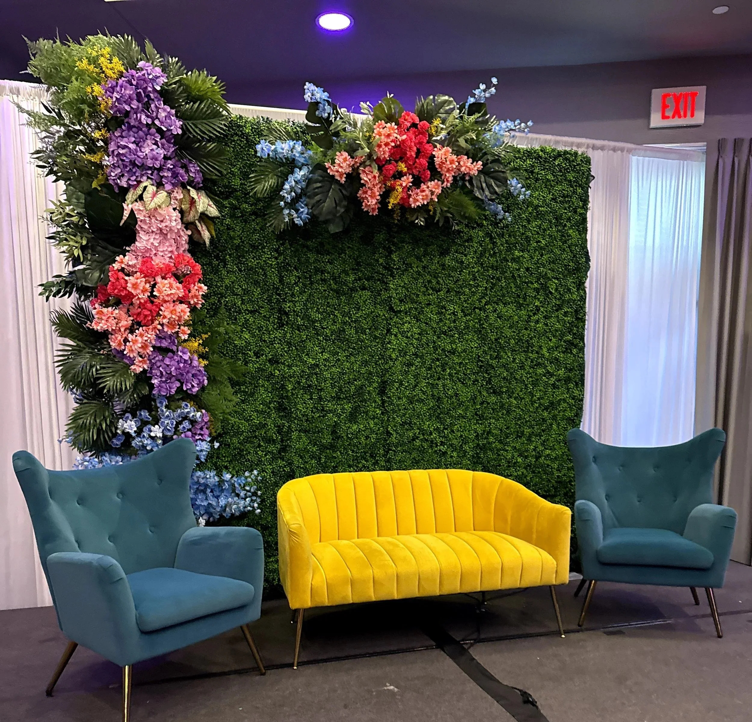 Colorful flower arrangements on a green hedge backdrop with purple ceiling lights and yellow and teal chairs in a modern indoor setting.