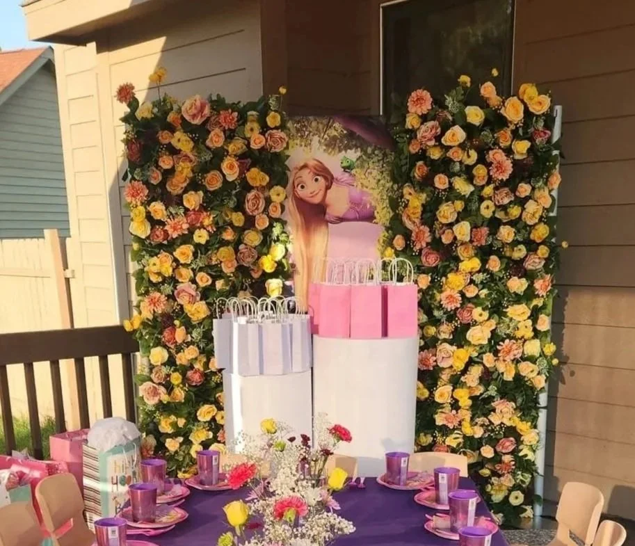 A birthday party setup outside with a Rapunzel theme, featuring a large flower wall, a Rapunzel poster, pink and purple tableware, and gift bags.