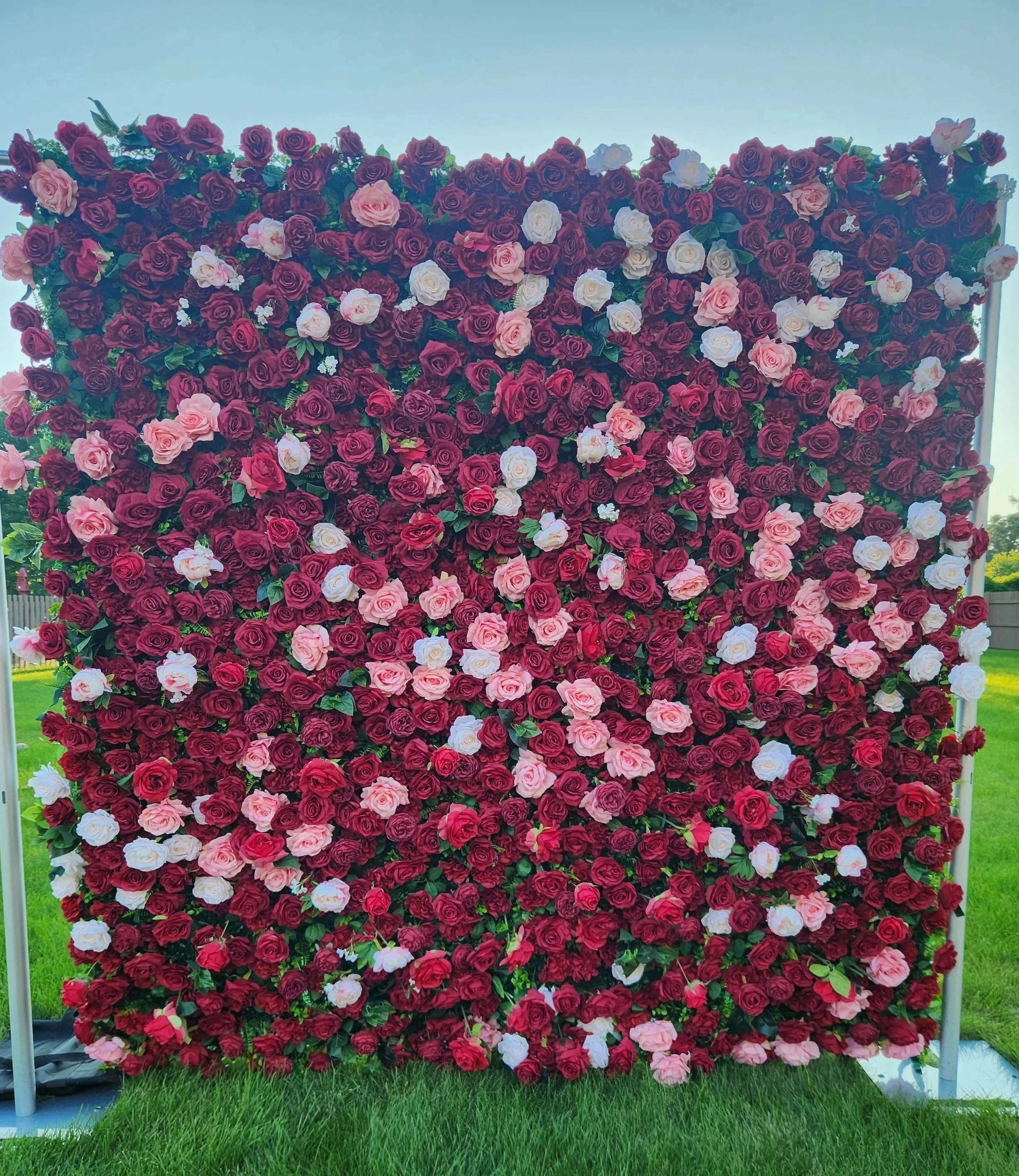 Large floral display with red, pink, and white roses, set outdoors on a metal frame with green grass in the foreground and blue sky in the background.