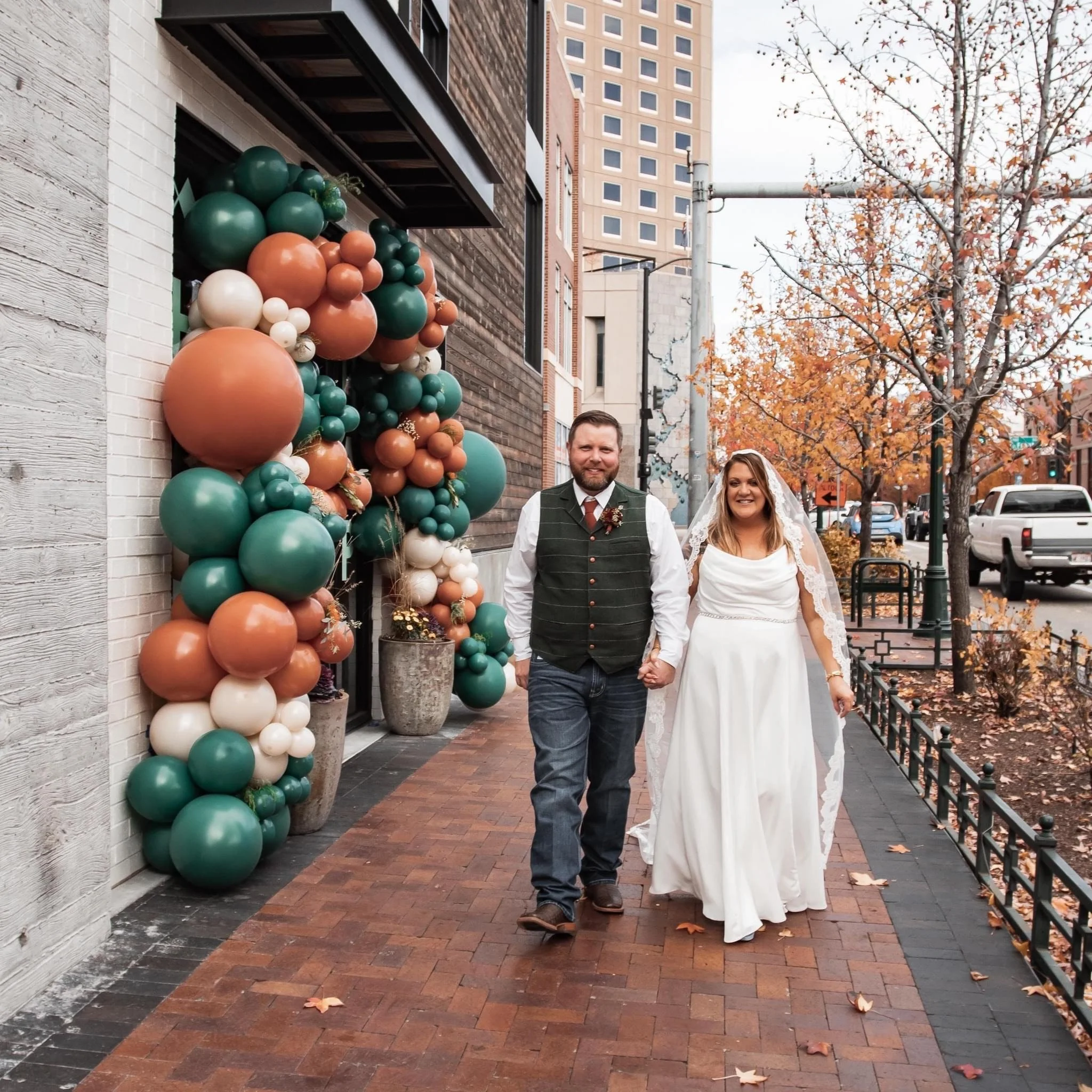 A newlywed couple walking hand in hand on a city sidewalk during fall, with colorful balloon decorations and autumn trees in the background.