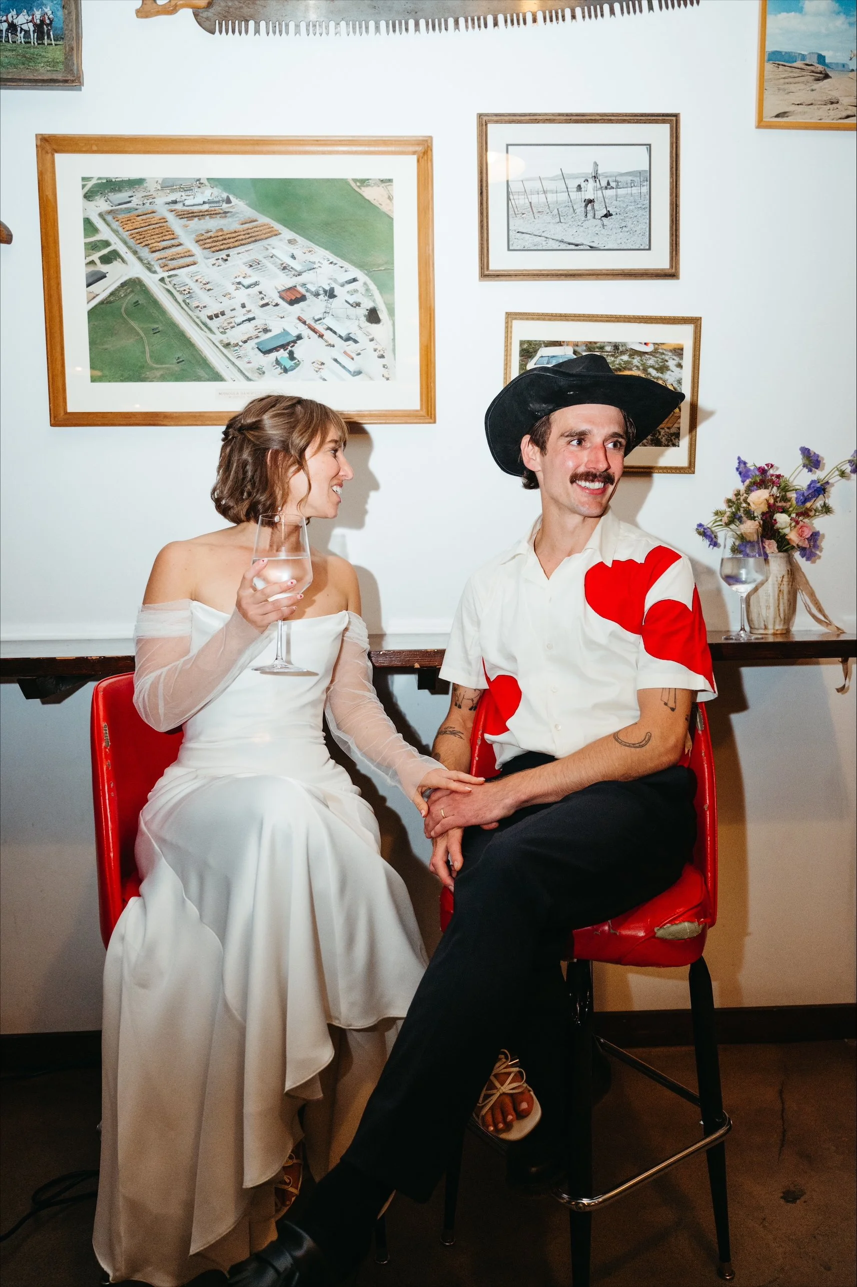A couple sitting on red chairs, with the woman in a white off-shoulder dress holding a glass, and the man in a cowboy hat and a white shirt with red hearts, holding hands and smiling, in a room with pictures and flowers on the wall.