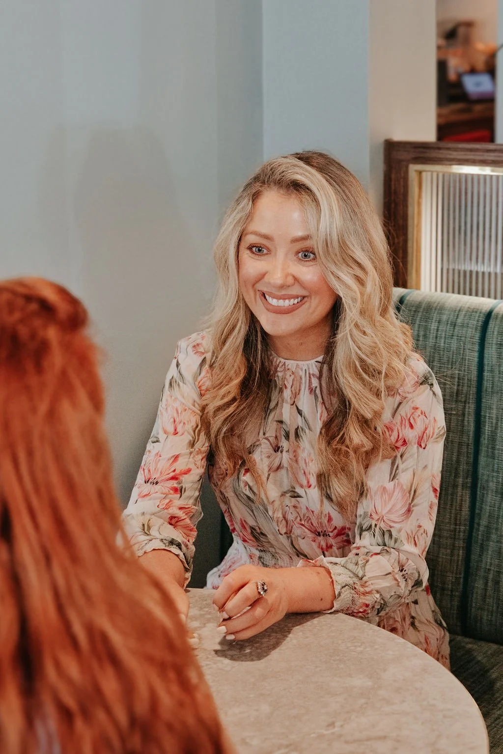 Long blonde curly haired woman wearing flowery dress sat speaking to someone