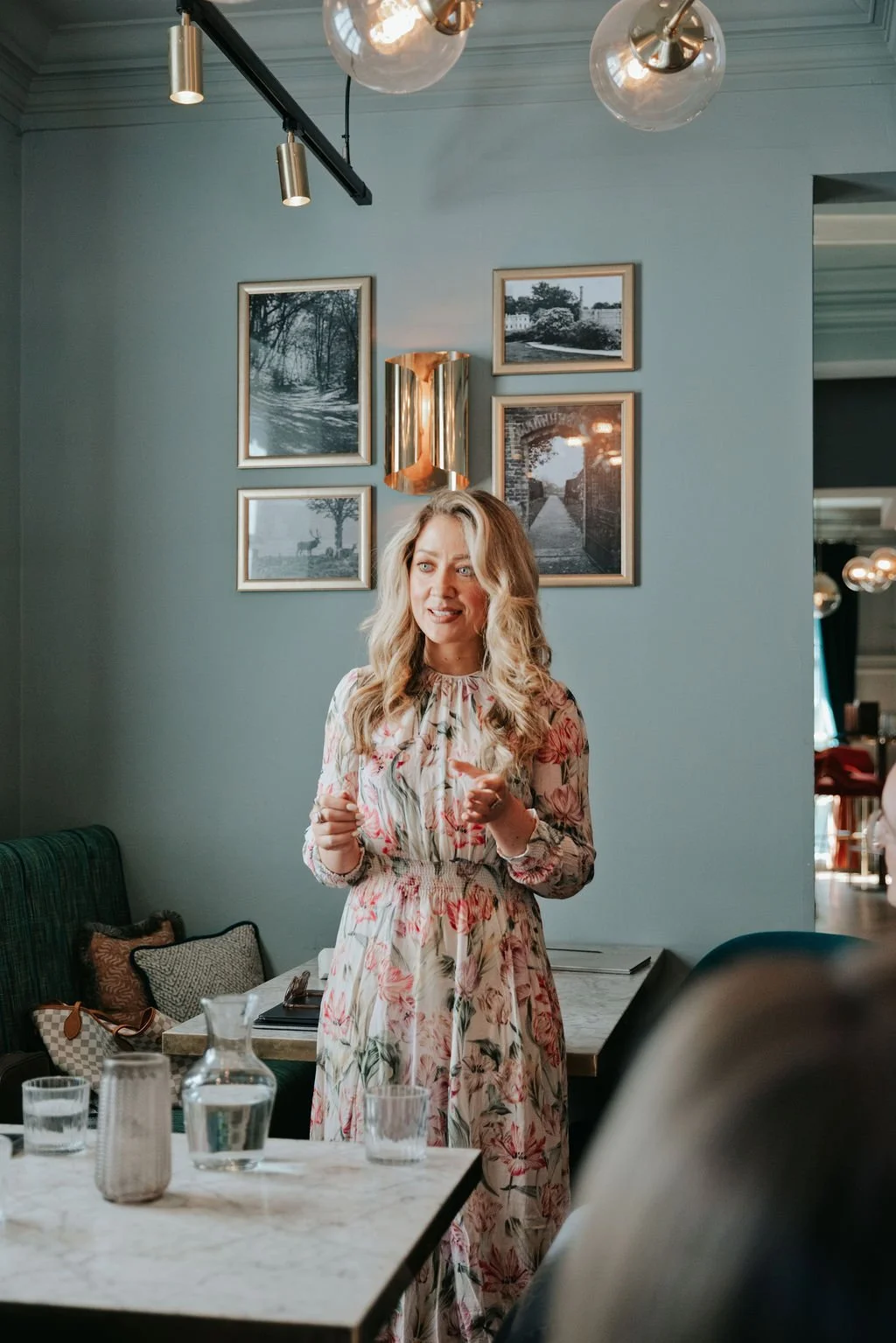 Woman with blonde curly hair wearing a flowery dress standing delivering a talk to a group