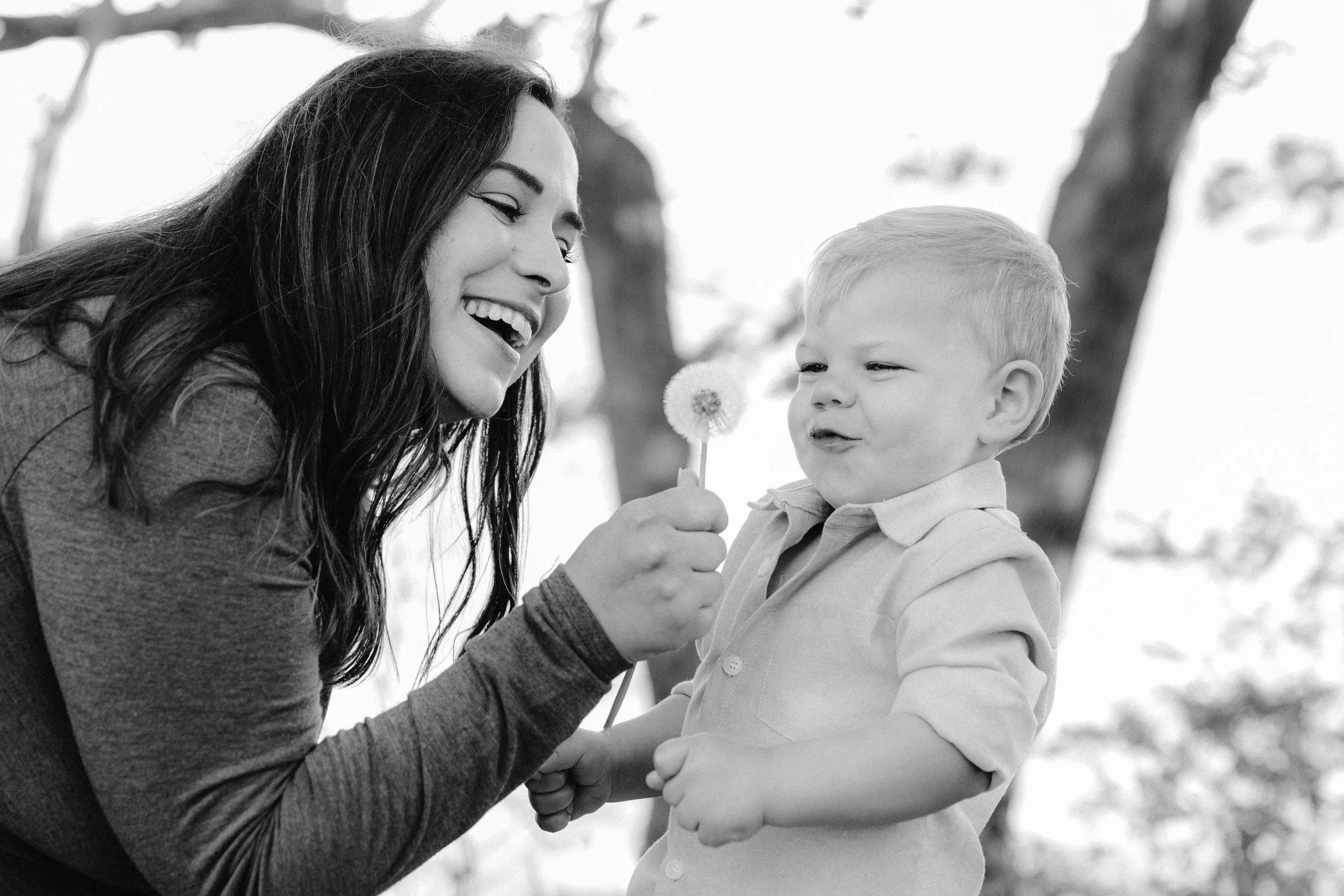  A black and white candid photo of a mother showing her young son how to blow a dandelion 