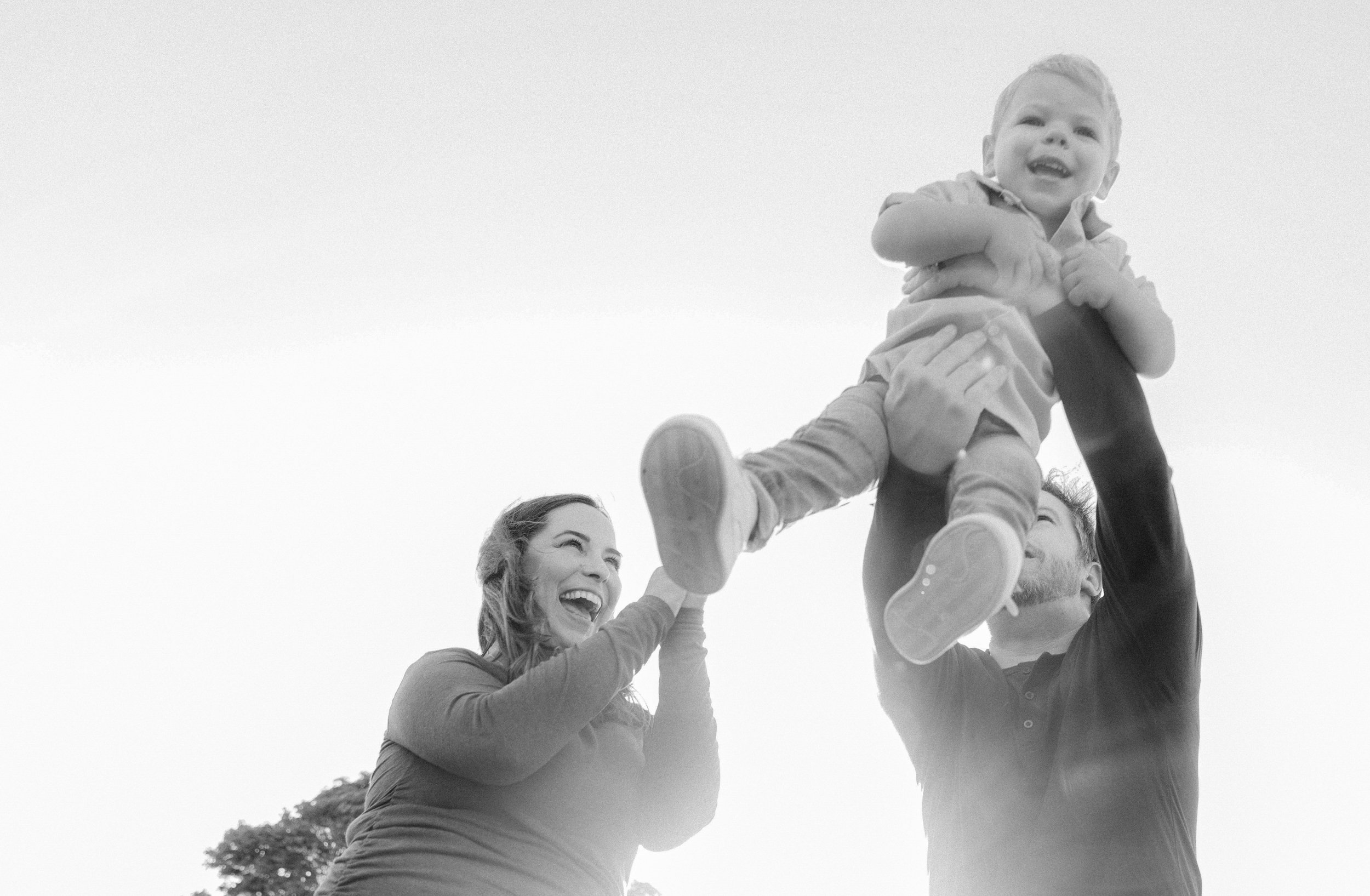  In this black and white image a mother and father swing their son up into the area as they are backlit by the sun 