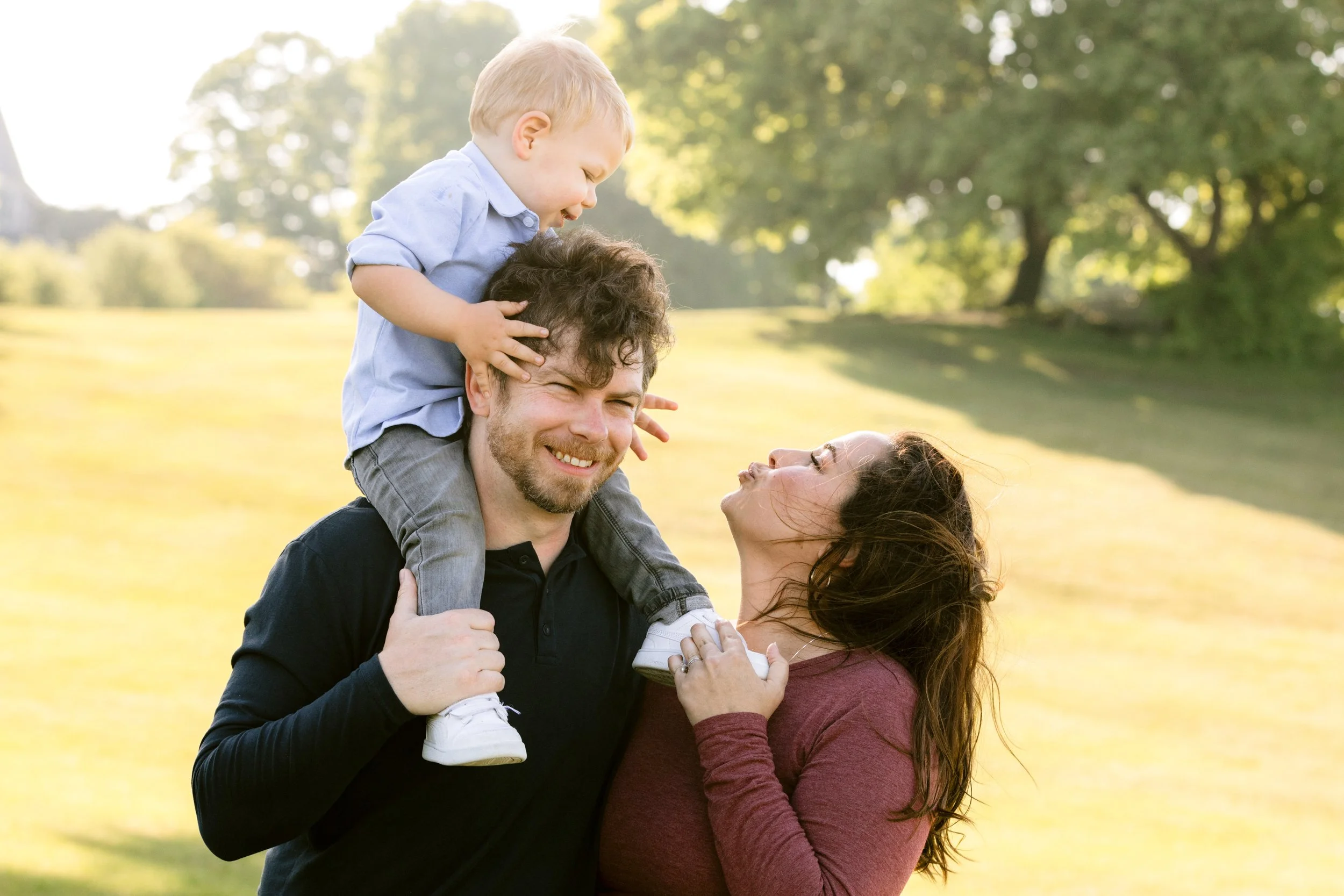  A father has his son on his shoulders with the mom making a kissing face up at him 